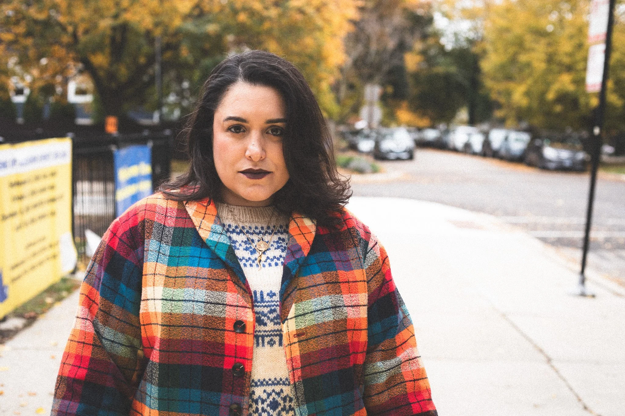 A young woman standing outdoors on a sidewalk during autumn, wearing a colorful plaid jacket and dark lipstick, with trees and parked cars in the background.