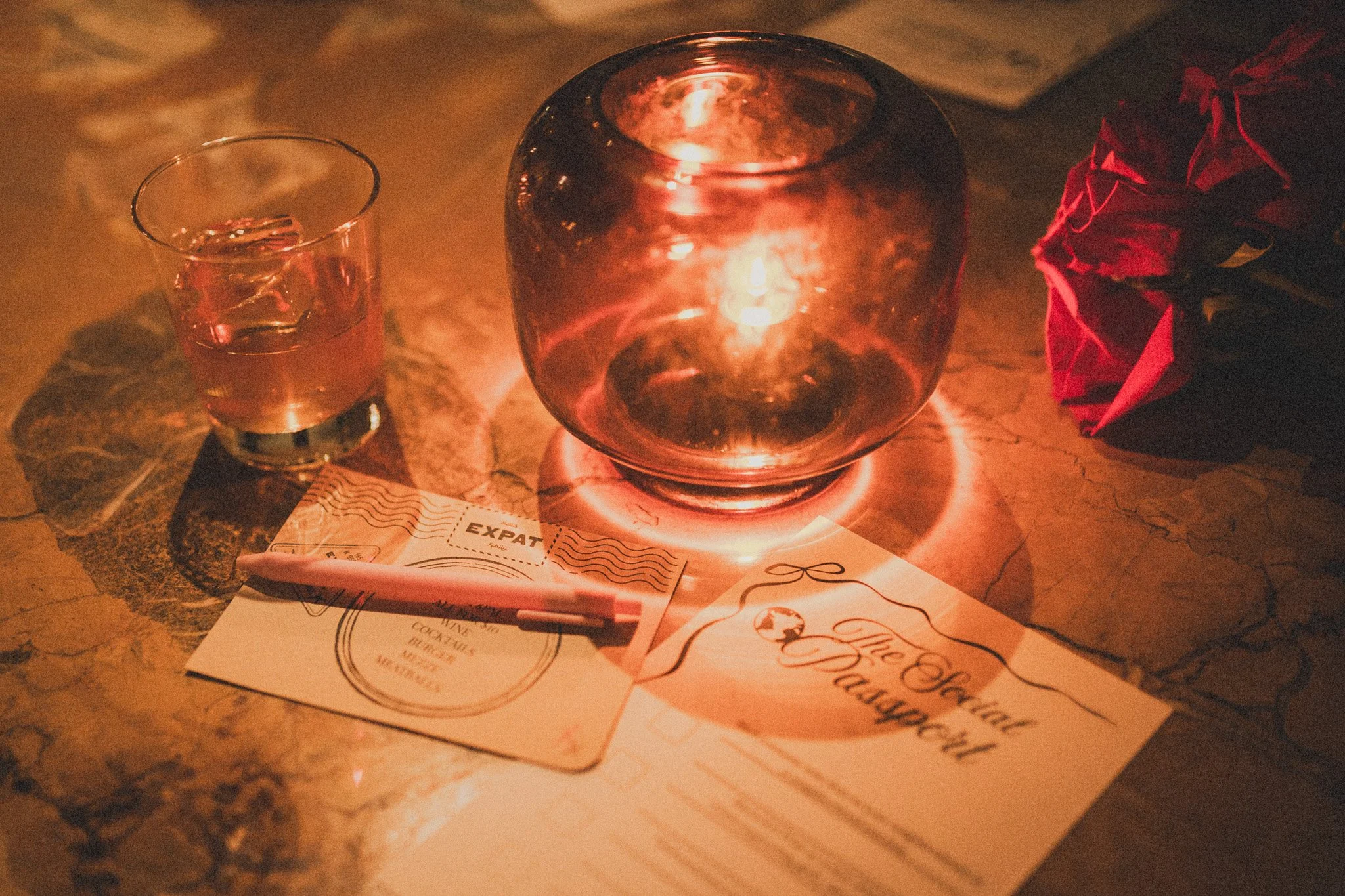 A candle inside a glass holder illuminating a table with a handwritten menu, a glass of water, and a red rose on the side.