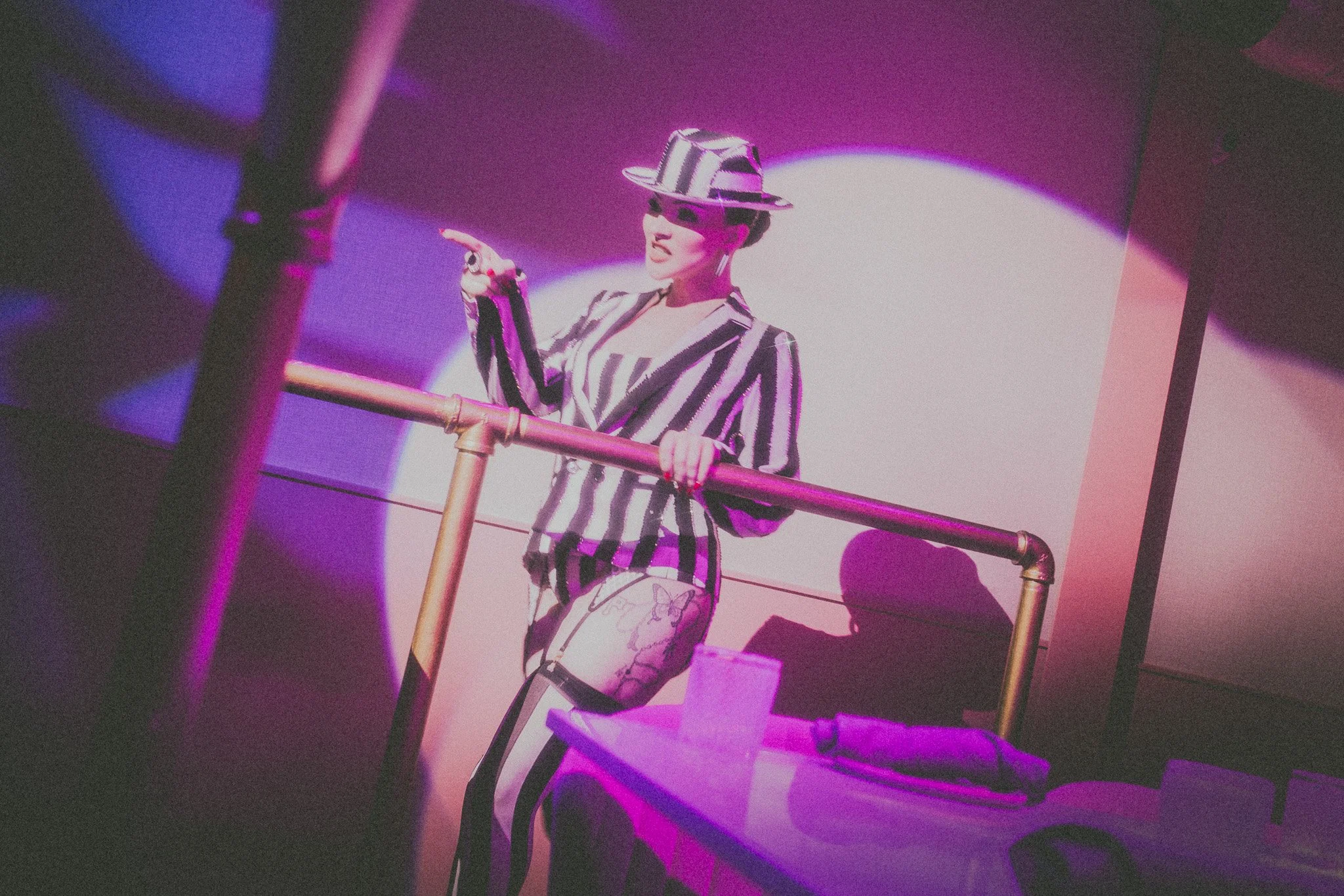 A person in a striped suit and hat standing on a platform with a pink and purple background and spotlight.