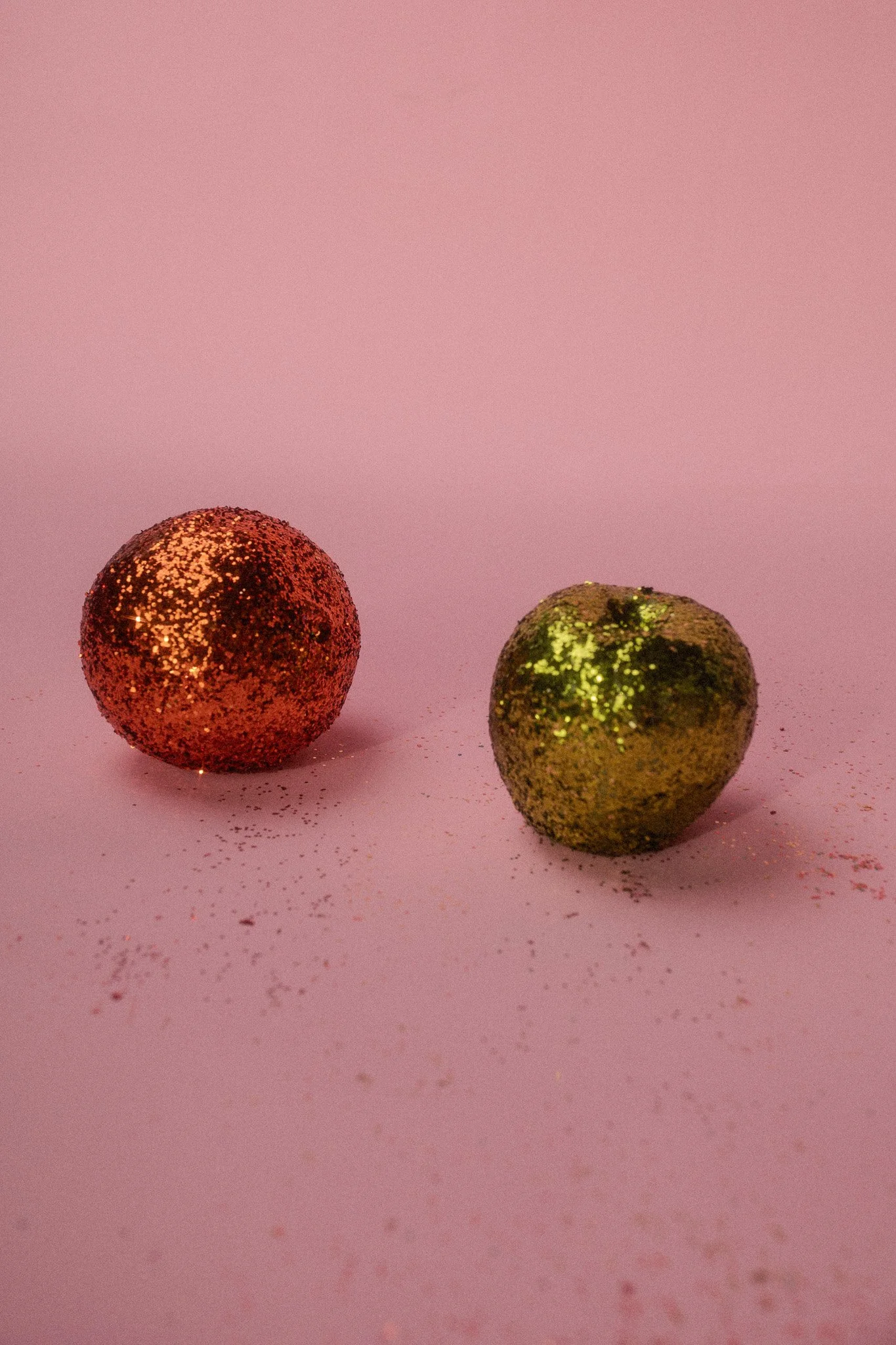 Red and gold glittered spherical and apple-shaped objects on pink surface with scattered glitter