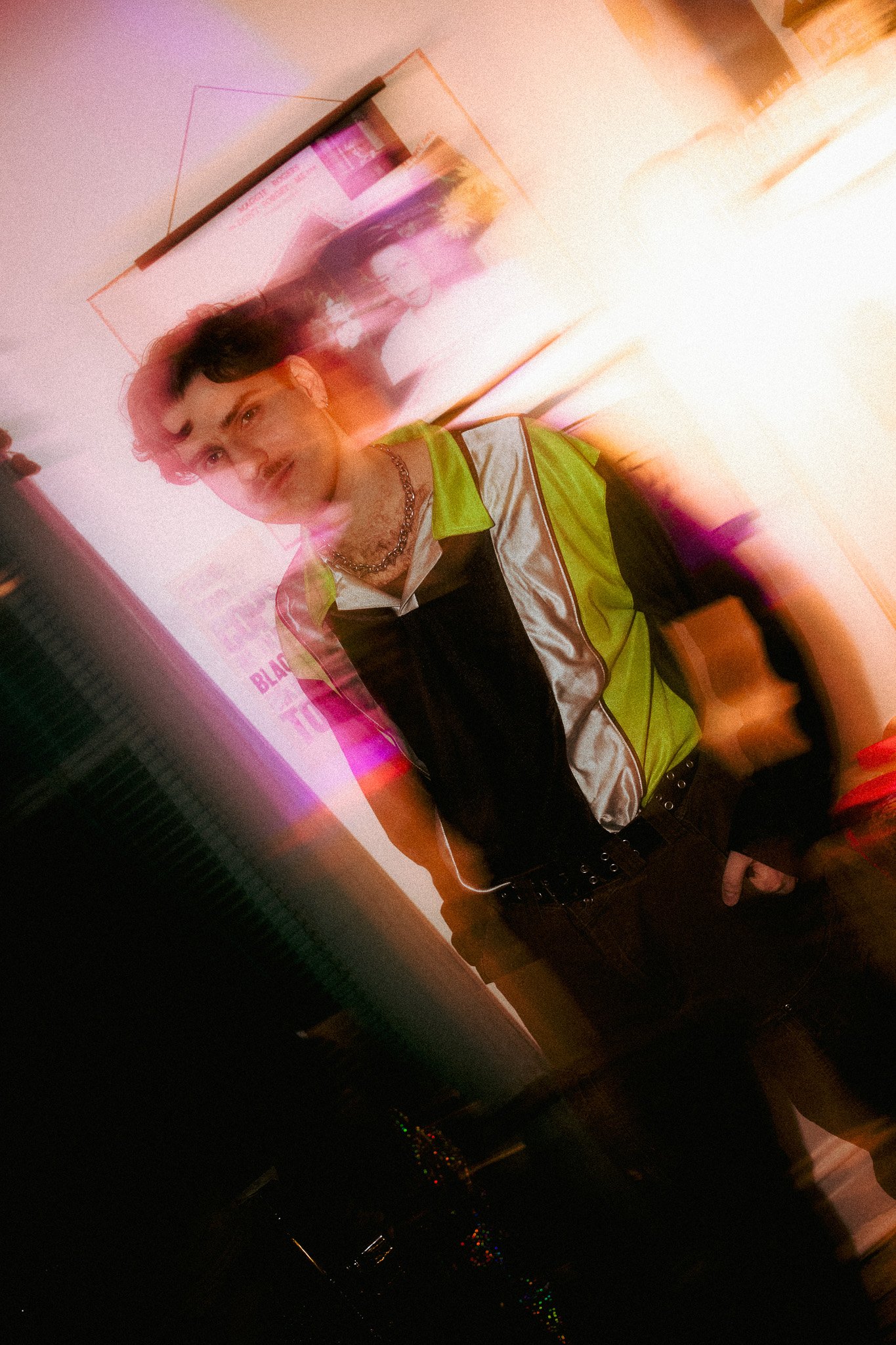 A young man with short, curly hair wearing a black and neon yellow vest over a white shirt, standing in front of a photo collage with a blurry background, and illuminated by colorful lights creating a dynamic, motion-blur effect.