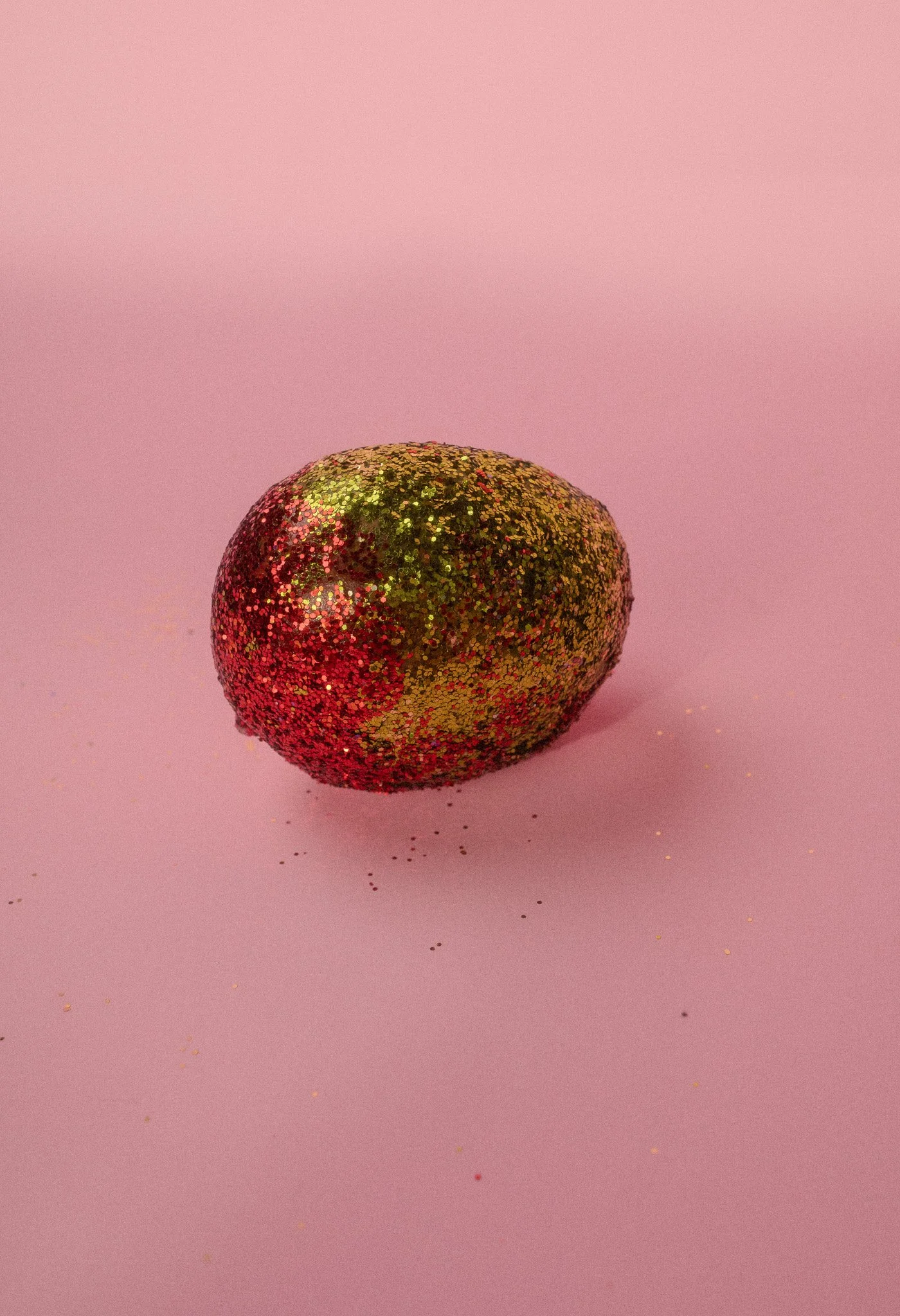 A small object resembling a lemon, covered in multicolored glitter, placed on a pink background.