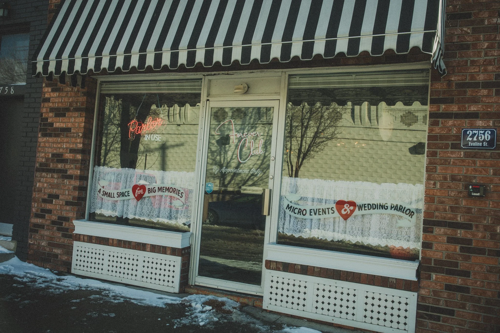The storefront of a small business with a black and white striped awning, brick exterior, and glass door. The windows have lace curtains and neon signs reading 'Pavilion' and 'Forever Club'. There are promotional signs on the windows advertising a sm