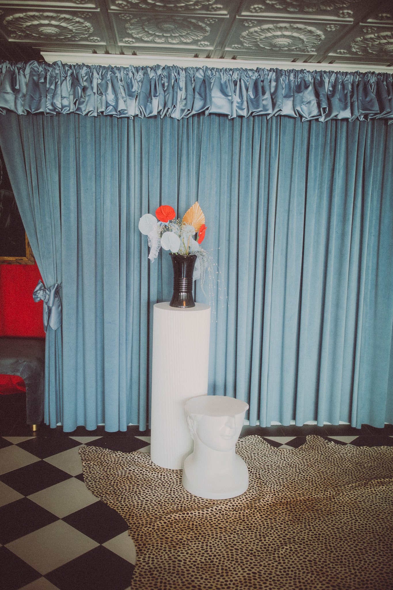 A decorative indoor setting featuring a tall black vase with red, white, and gold paper flowers, placed on white cylindrical pedestal and a white ceramic bust-shaped container, against a backdrop of closed blue curtains, with a black and white checke