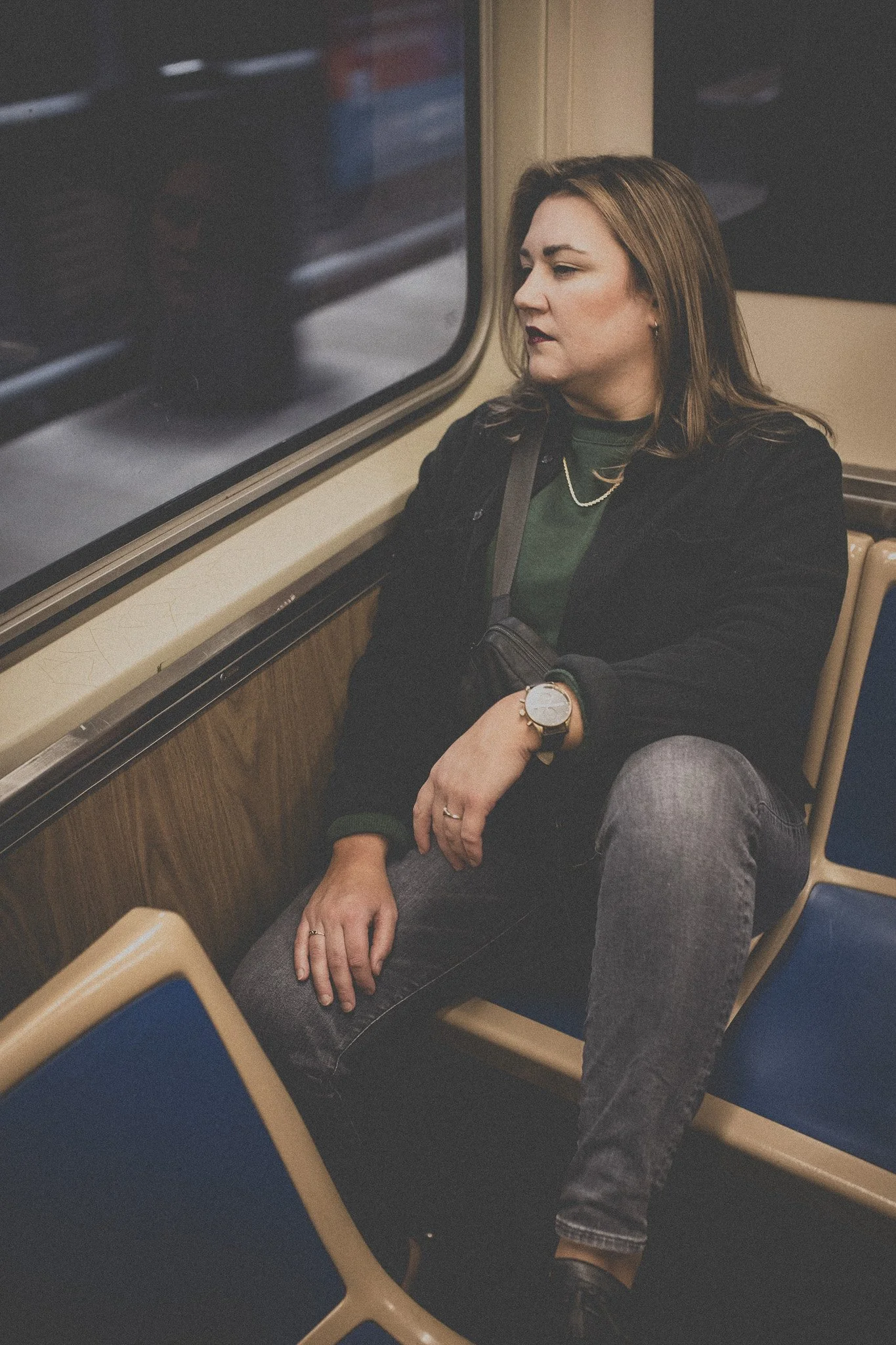 A woman sitting alone on a train, looking out the window with a thoughtful expression, wearing a black jacket, green shirt, jeans, and accessorized with a watch, rings, and a chain necklace.