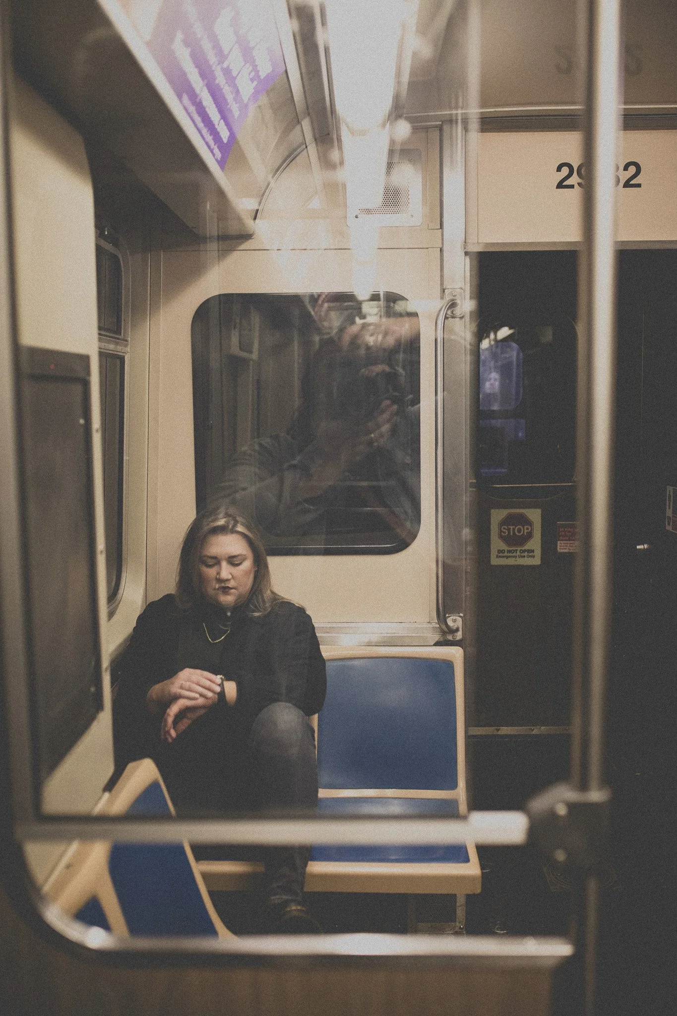 A woman sitting alone on a subway train, looking at her watch. The photo is reflected in the window, with the photographer visible in the reflection.