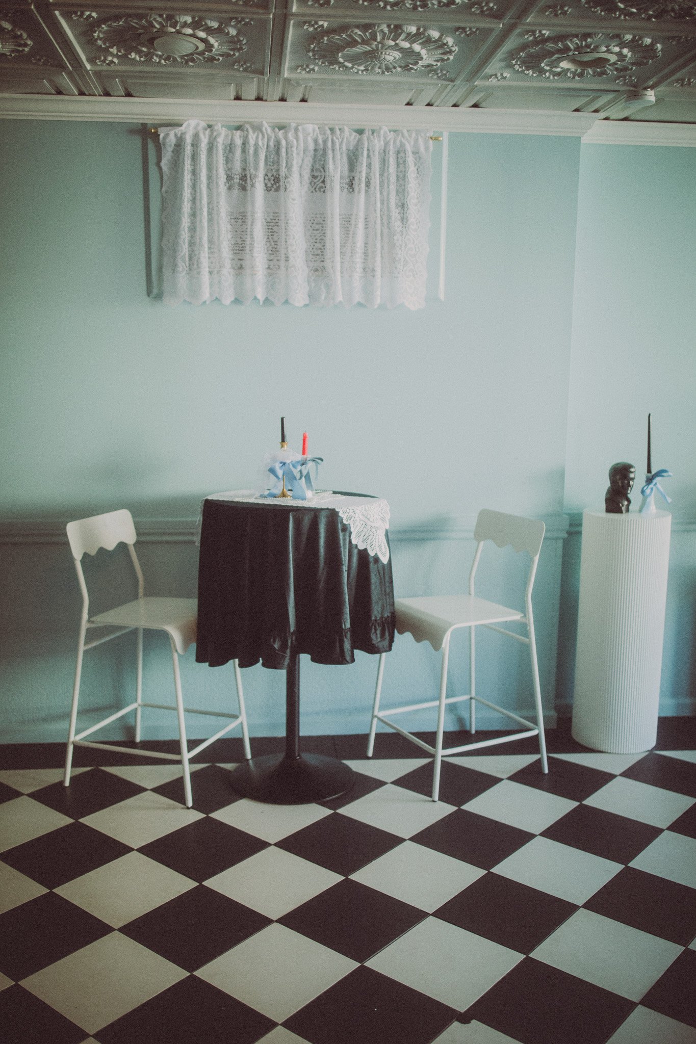 A small round table with a black tablecloth and a white lace overlay, with two white chairs on either side. There are two unlit candles in holders on the table, and a white curtain with lace hangs on the wall behind it. To the right, a white pedestal