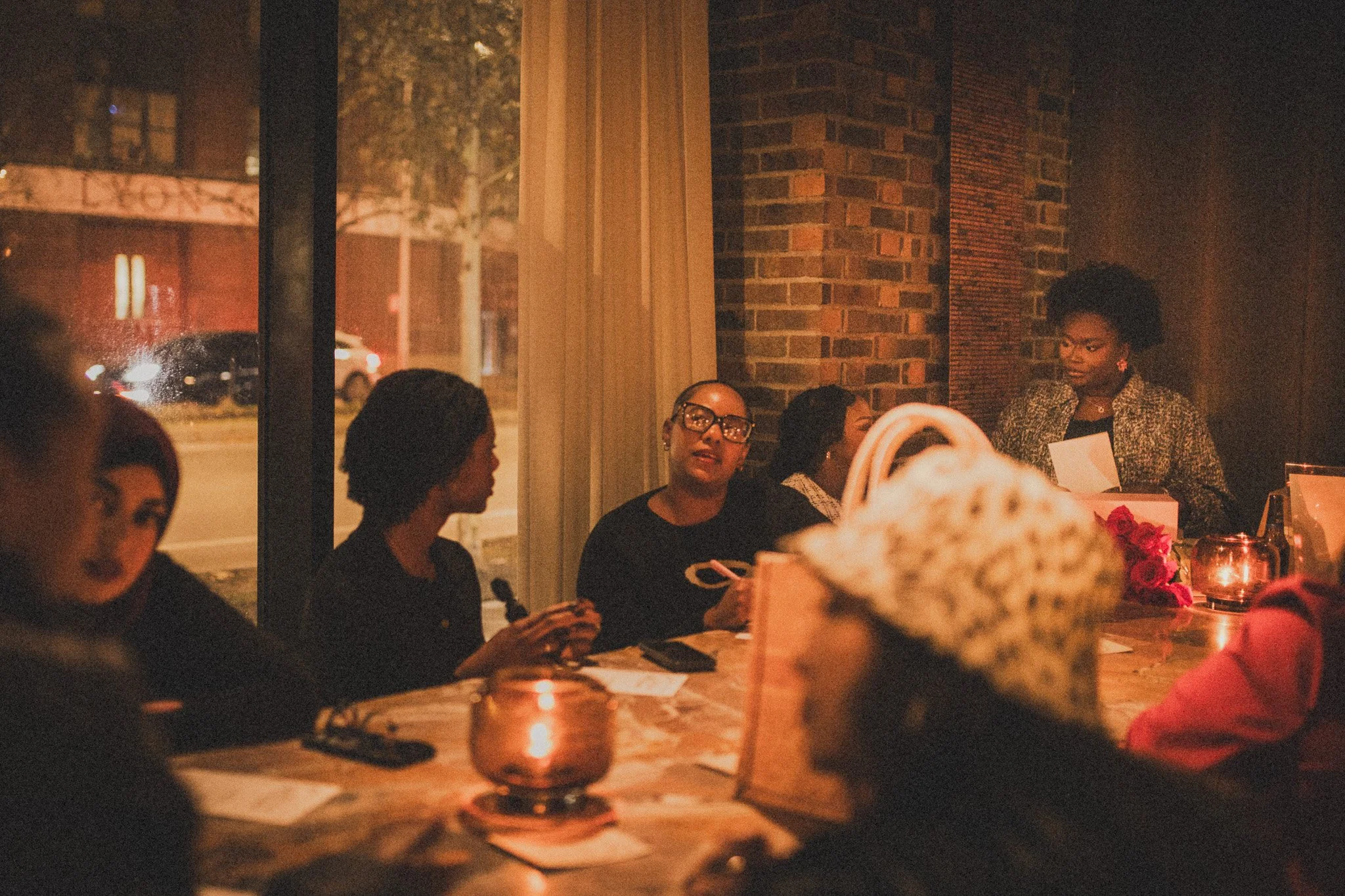 A group of women sitting at a table in a dimly lit restaurant or cafe, with candles providing warm lighting. Through a large window, a street scene with cars and trees is visible outside. One woman on the right side who is standing appears to be spea