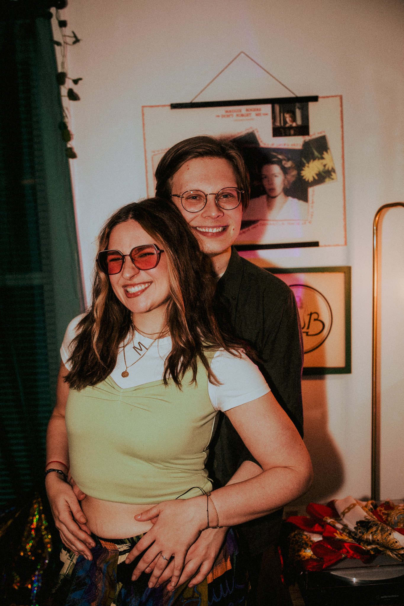 A smiling young woman with long wavy hair, wearing pink sunglasses, a white t-shirt, and a yellow top, is hugging a young man with short brown hair and glasses, standing in a room with colorful decorations and framed pictures on the wall.