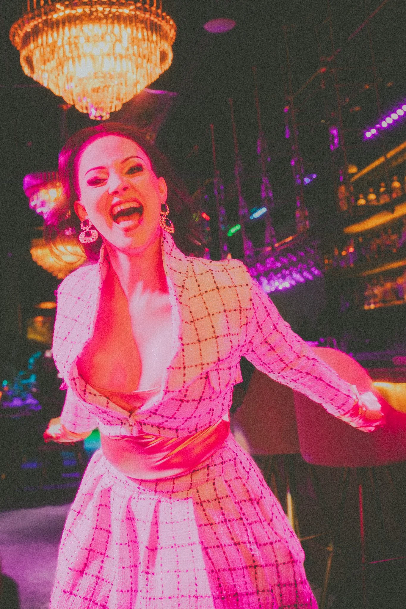 A woman with dark hair styled in an updo, wearing a pink and white checkered dress with a deep neckline, standing in a dimly lit room with vibrant colorful lighting and chandeliers in the background, smiling energetically.