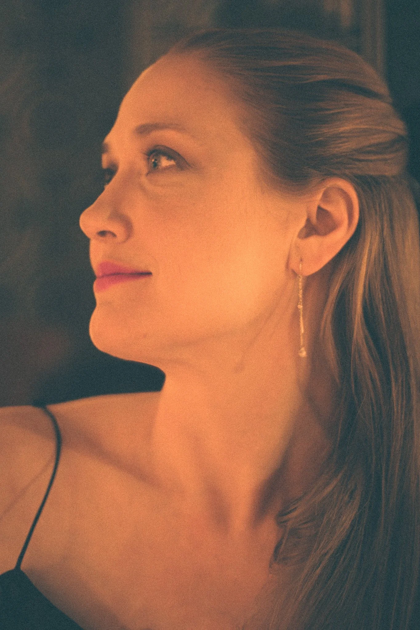 Close-up profile of a woman with long, light brown hair, wearing earrings and a dark strap top, looking away with a slight smile.