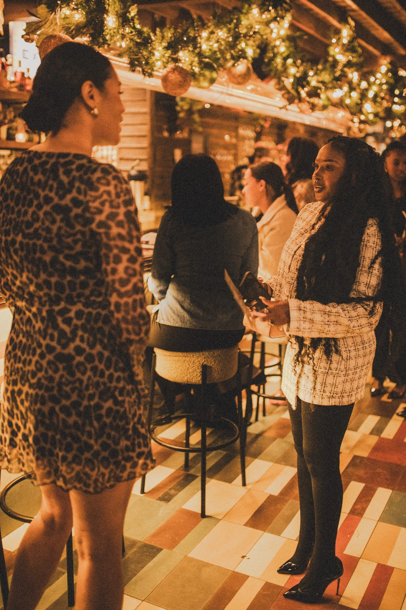 Women chatting in a warmly lit, outdoor holiday-themed setting with Christmas lights and decorations.