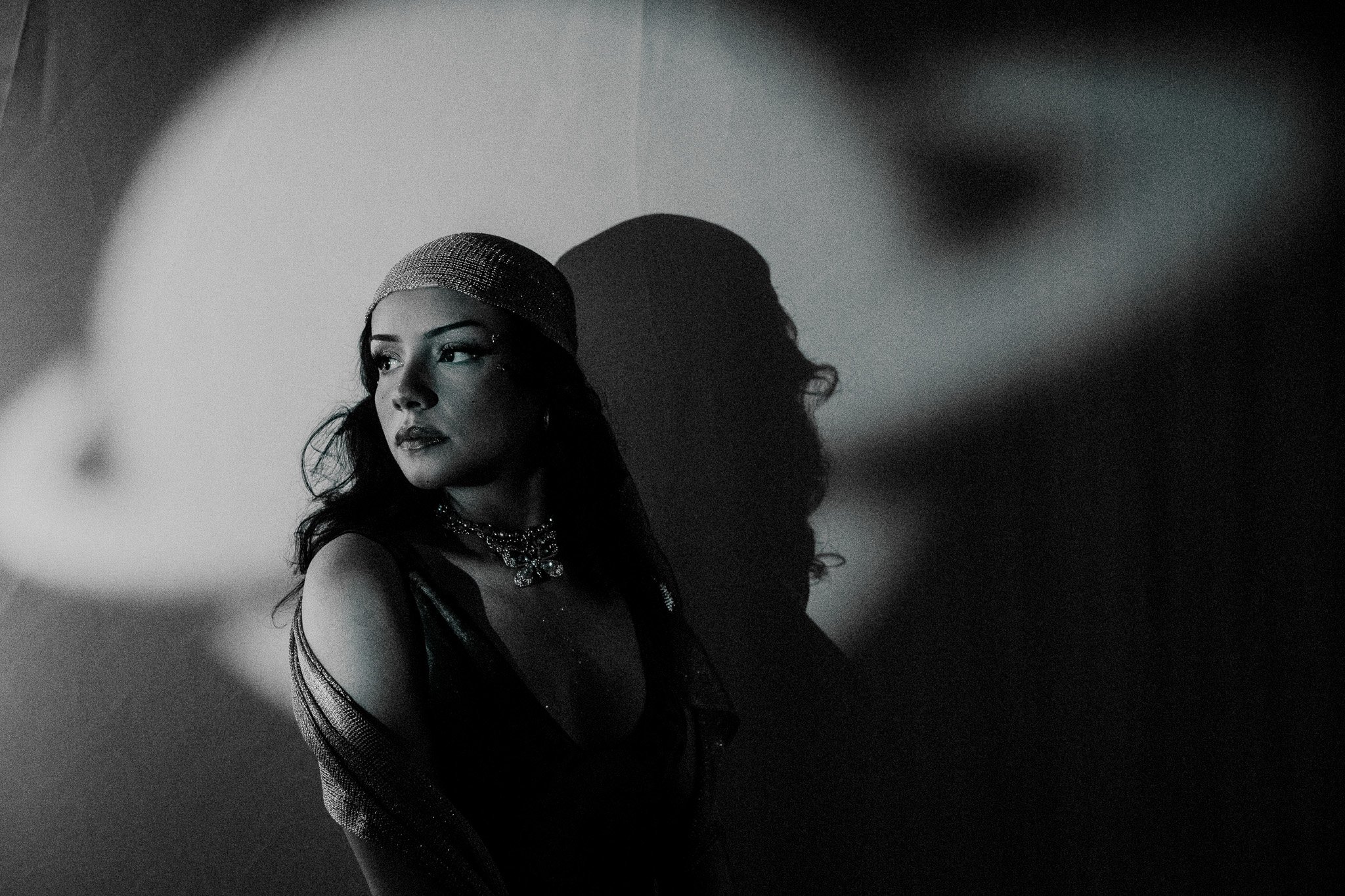 Black and white photo of a woman with dark wavy hair, wearing a sparkly headband and necklace, looking to her left, casting a shadow on the wall behind her.