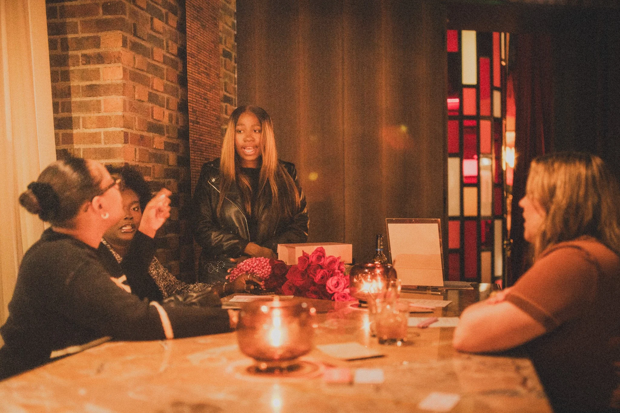A woman standing and speaking to three women sitting around a table in a dimly-lit room with brick walls and colorful stained glass window. The table has candles, flowers, and papers on it.