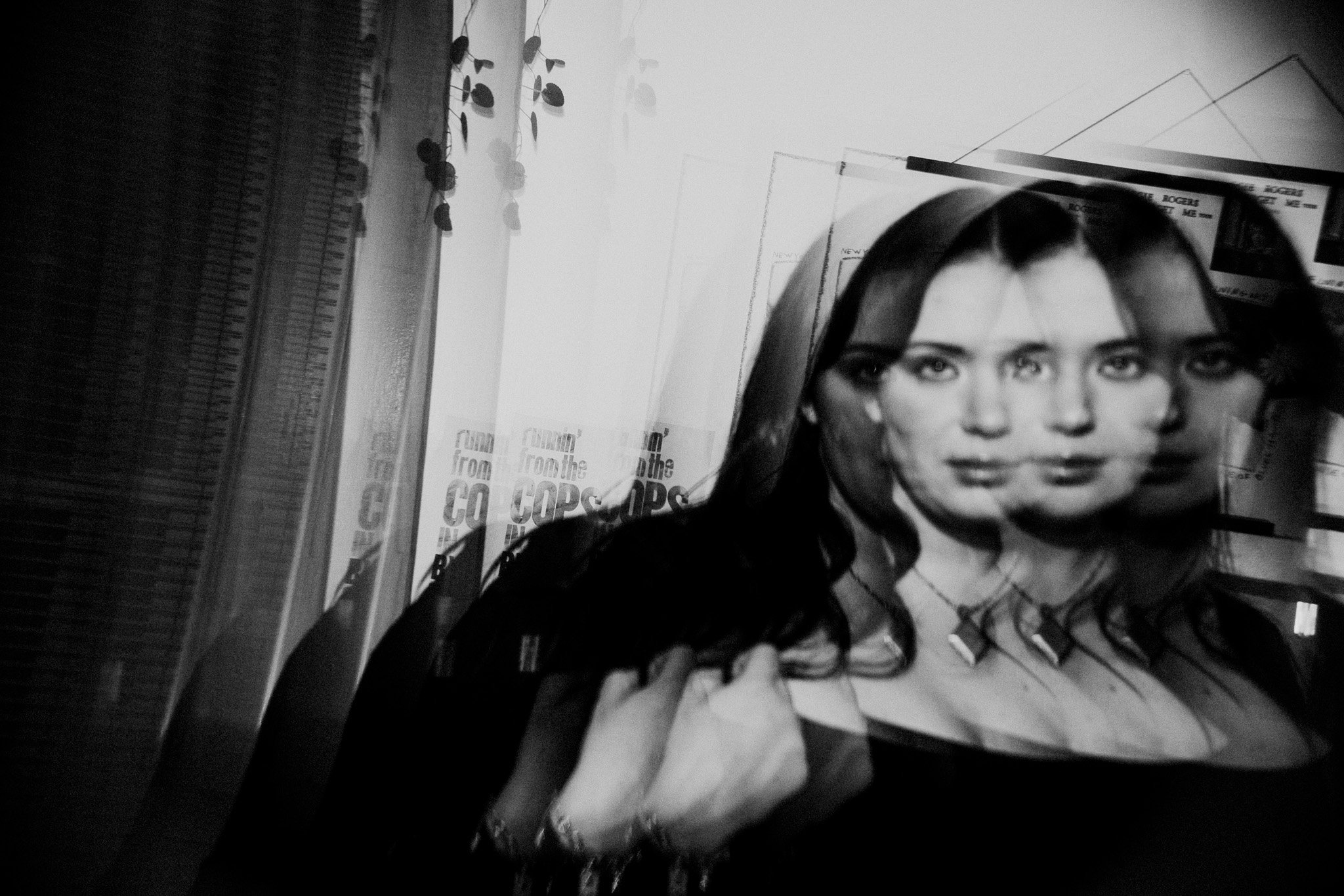 Black and white photo of a woman with long dark hair, multiple overlapping images creating a distorted effect, with posters in the background.