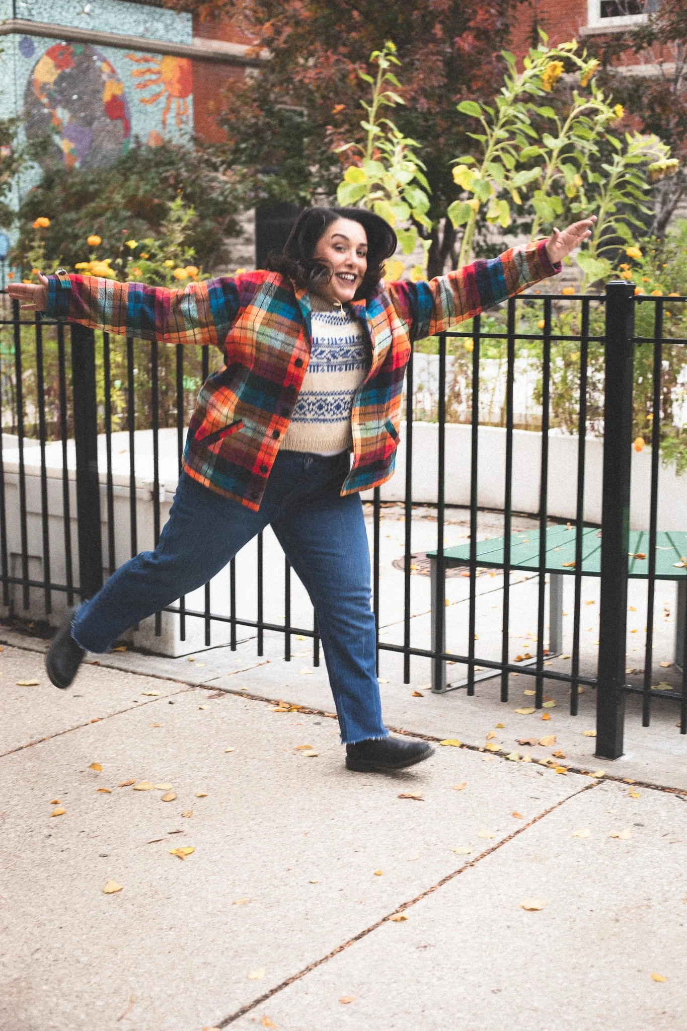 A woman with brown hair wearing a multicolored plaid jacket, sweater, and blue jeans walking outdoors with arms outstretched and smiling.