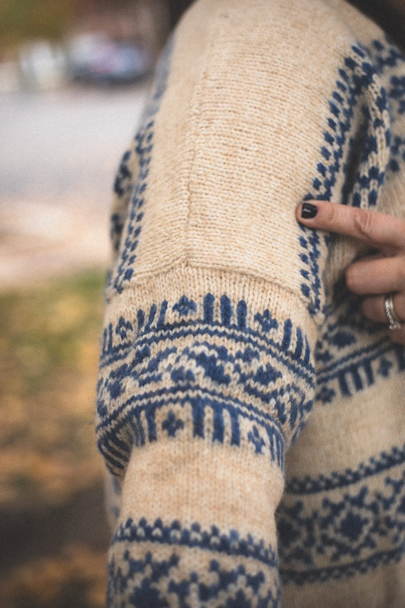 Close-up of a knit sweater with a blue and beige pattern on the sleeve, with a person's hand pointing at the sleeve.