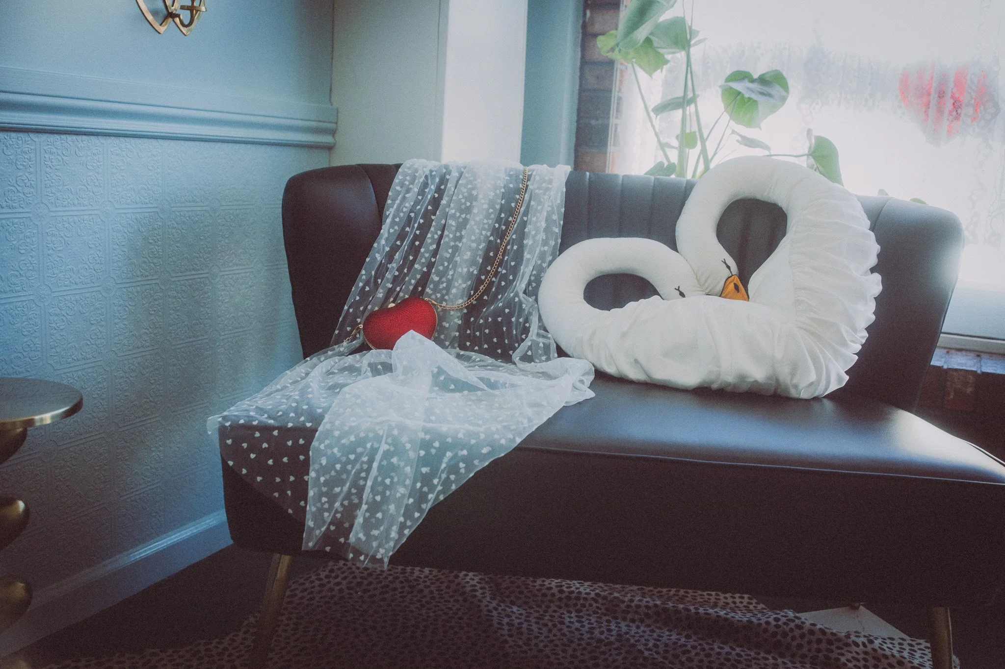 Black leather sofa decorated with two white swan-shaped pillow and sheer polka-dotted white fabric, in front of a window with a green plant on the sill, and a red heart-shaped decoration with a gold chain.
