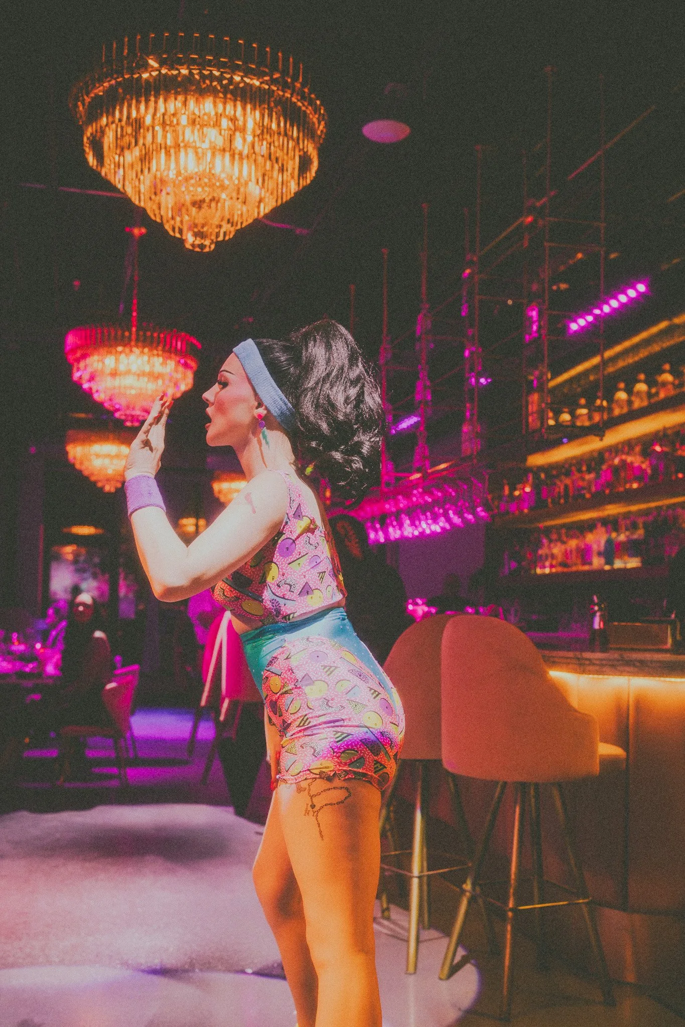 A woman with dark, wavy hair in a retro outfit, in a nightclub with colorful lighting, chandeliers, and a bar in the background.
