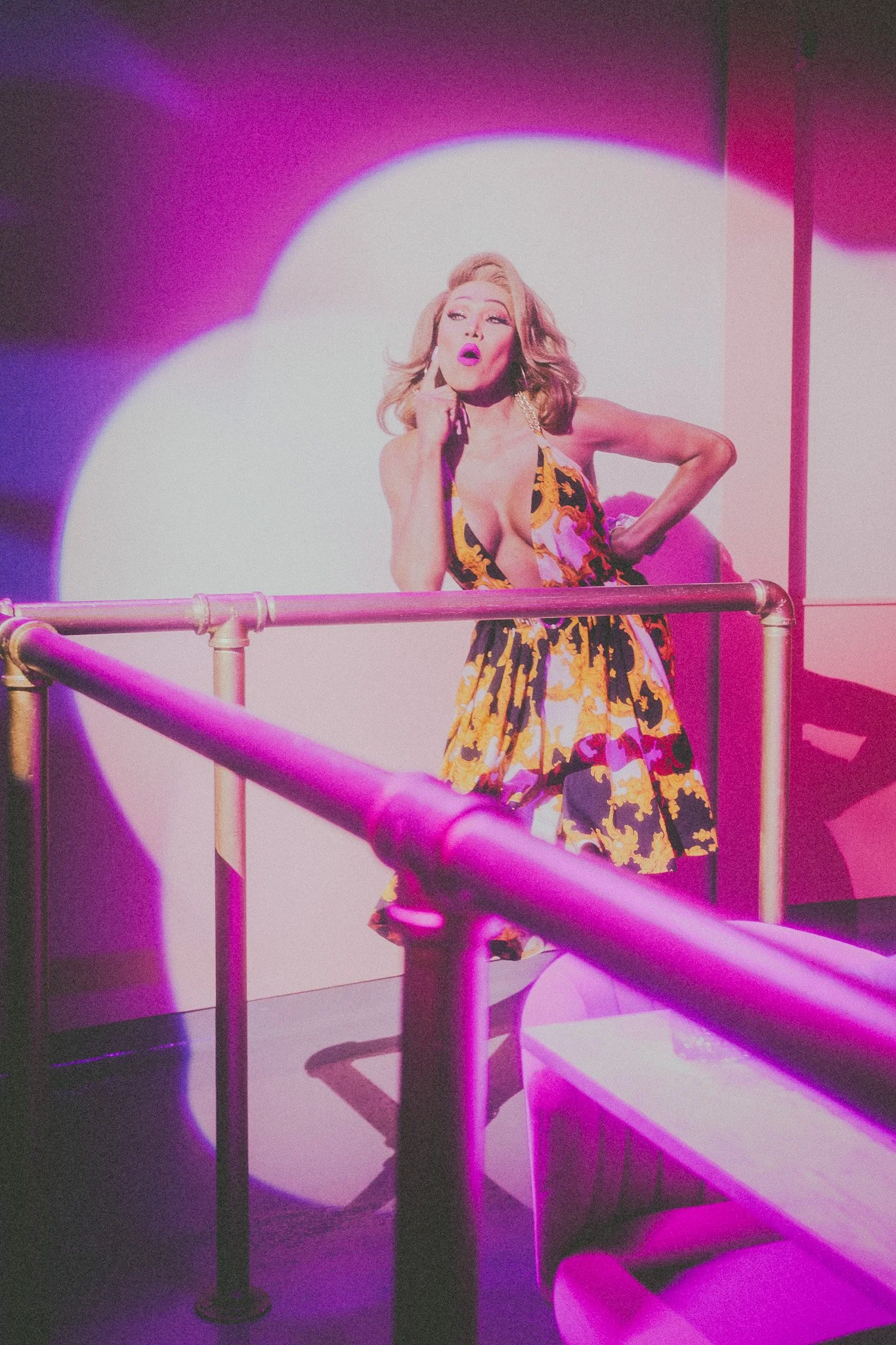 A woman wearing a colorful dress with a plunging neckline is standing behind a metal railing, striking a pose with one hand on her chin and the other on her hip, in front of a lit backdrop with purple and white lighting.