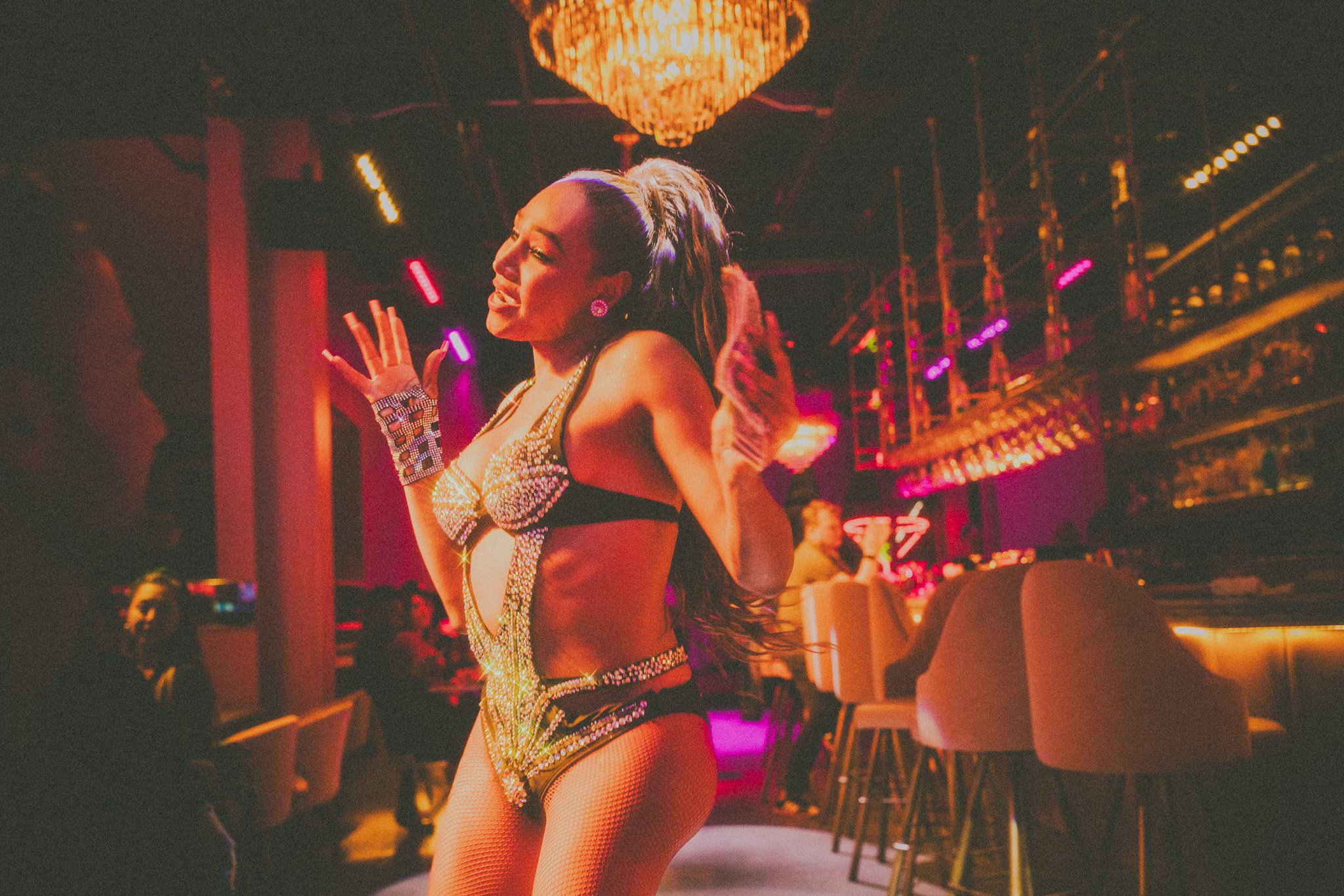 A woman performing a dance on a stage in a nightclub with colorful lighting, dressed in a sparkling, revealing costume with fishnet stockings, earrings, and gloves, with an audience and bar visible in the background.