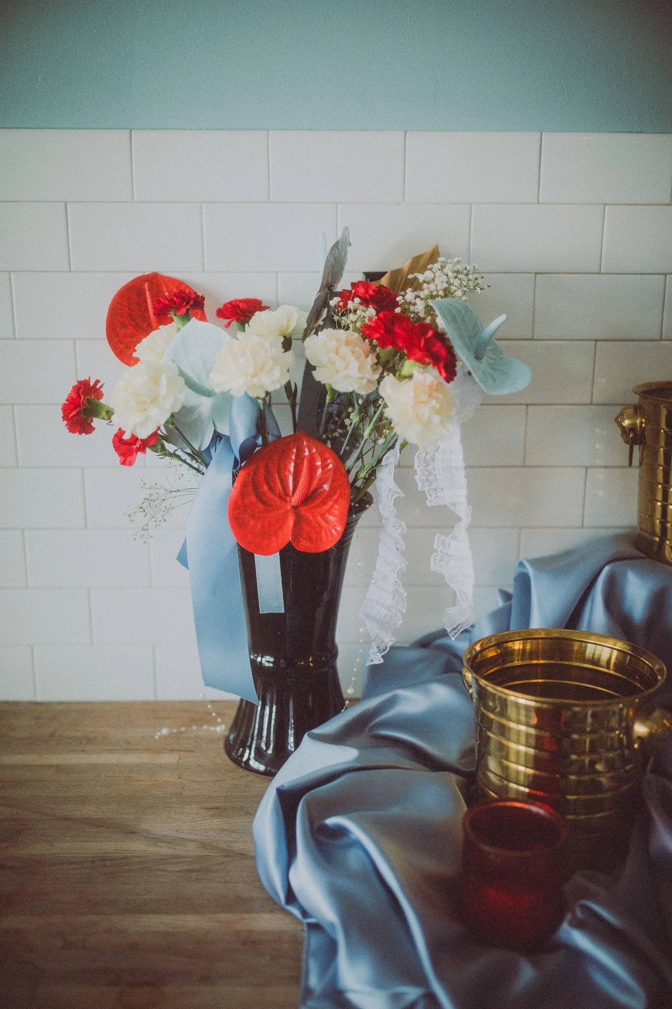 A floral arrangement of red, white, and pink flowers with large red anthuriums, set in a black vase, decorated with ribbons, placed on a wooden surface with golden vases and blue fabric nearby.