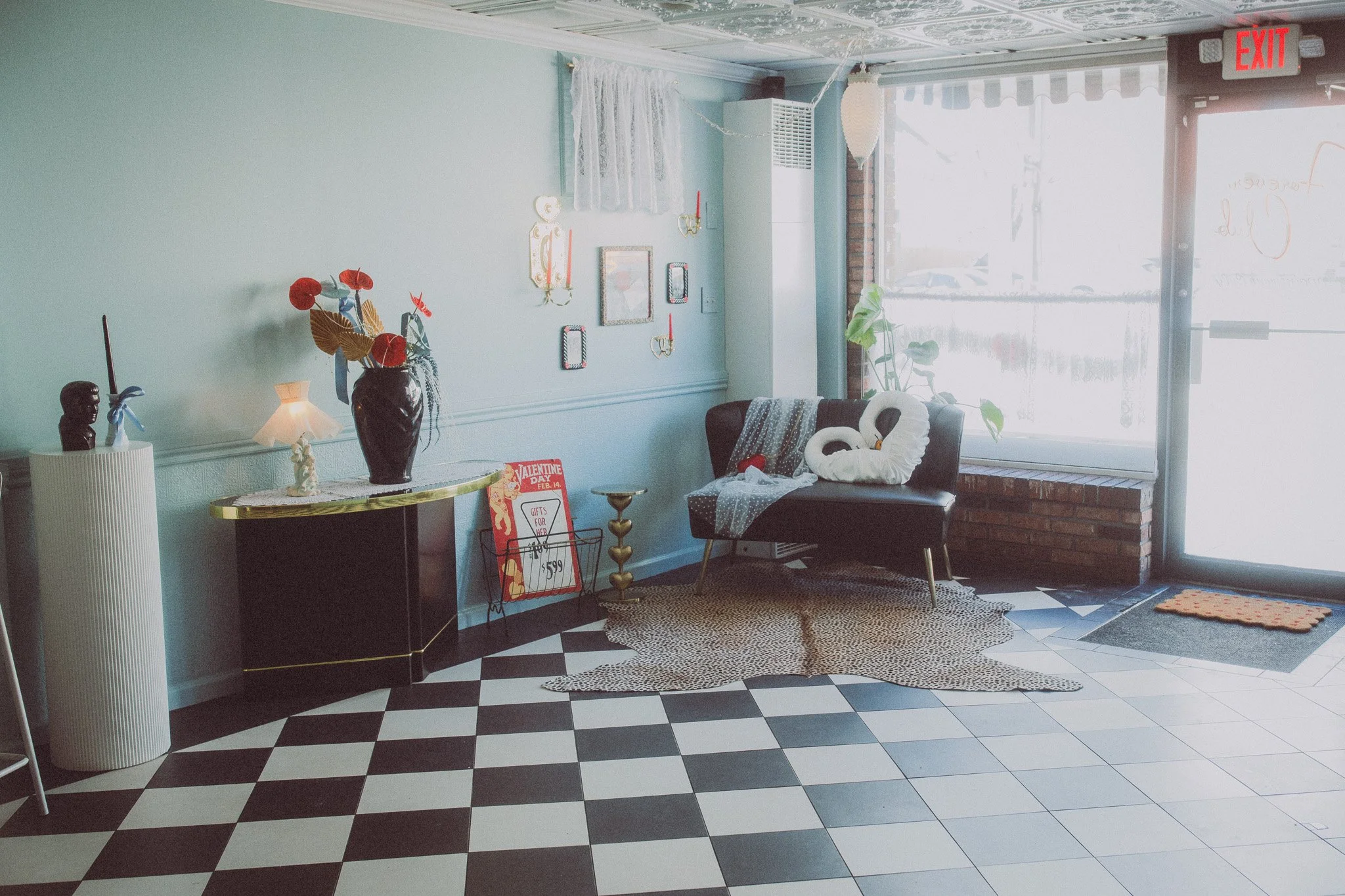 Interior of a cozy vintage store with checkered black and white floor, a black sofa with swan-shaped pillows, a leopard print rug, decorative wall art, a side table with a vase of flowers, and large windows letting in natural light.