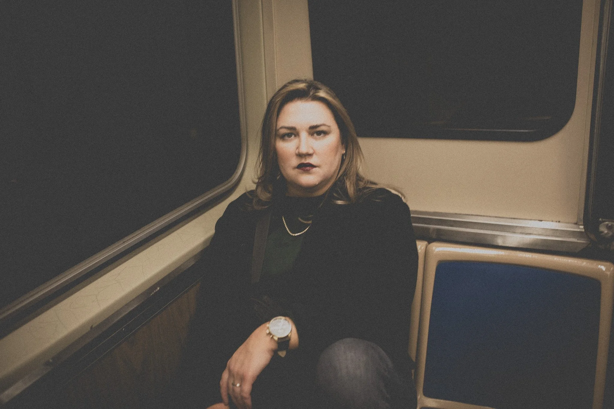 A woman with shoulder-length hair, dark lipstick, wearing a black jacket and a watch, sitting on a train at night.
