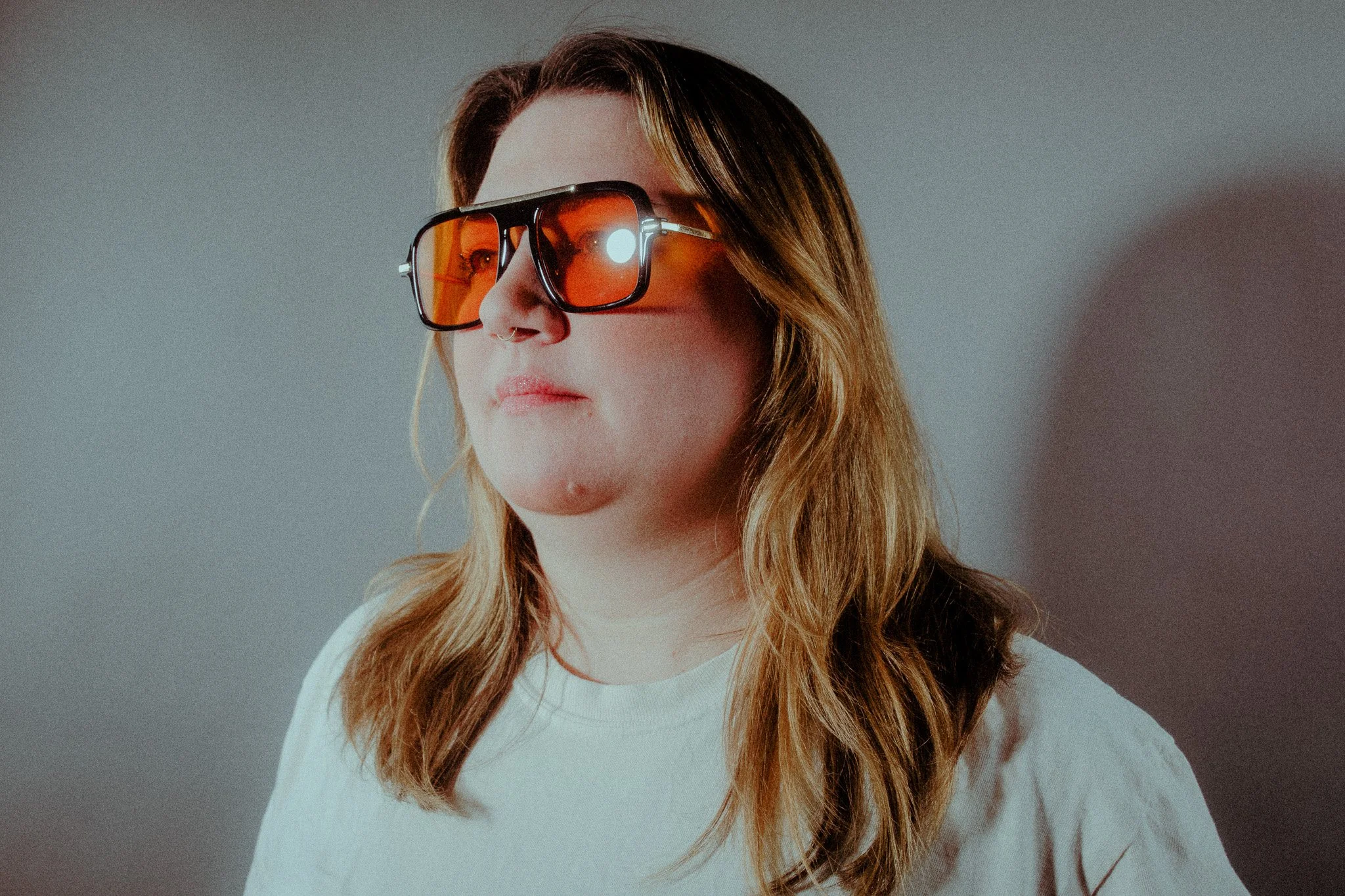 A woman with long brown hair wearing large orange-tinted sunglasses and a white shirt, standing against a neutral gray background.