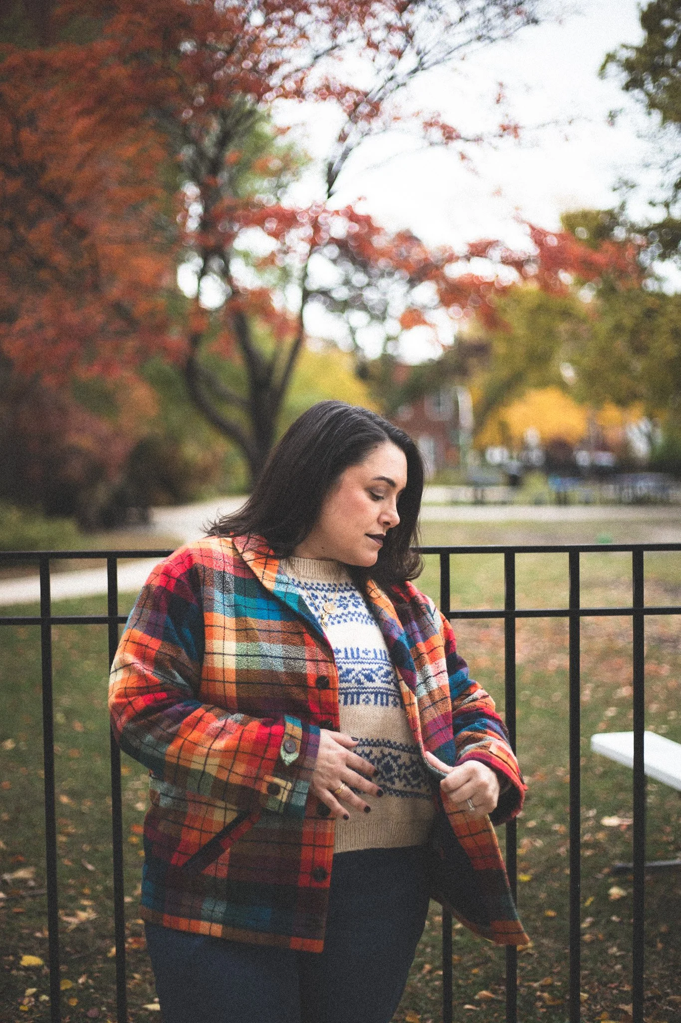 Woman with dark hair wearing a colorful plaid jacket and a patterned sweater, standing outdoors by a black fence with autumn trees in the background.