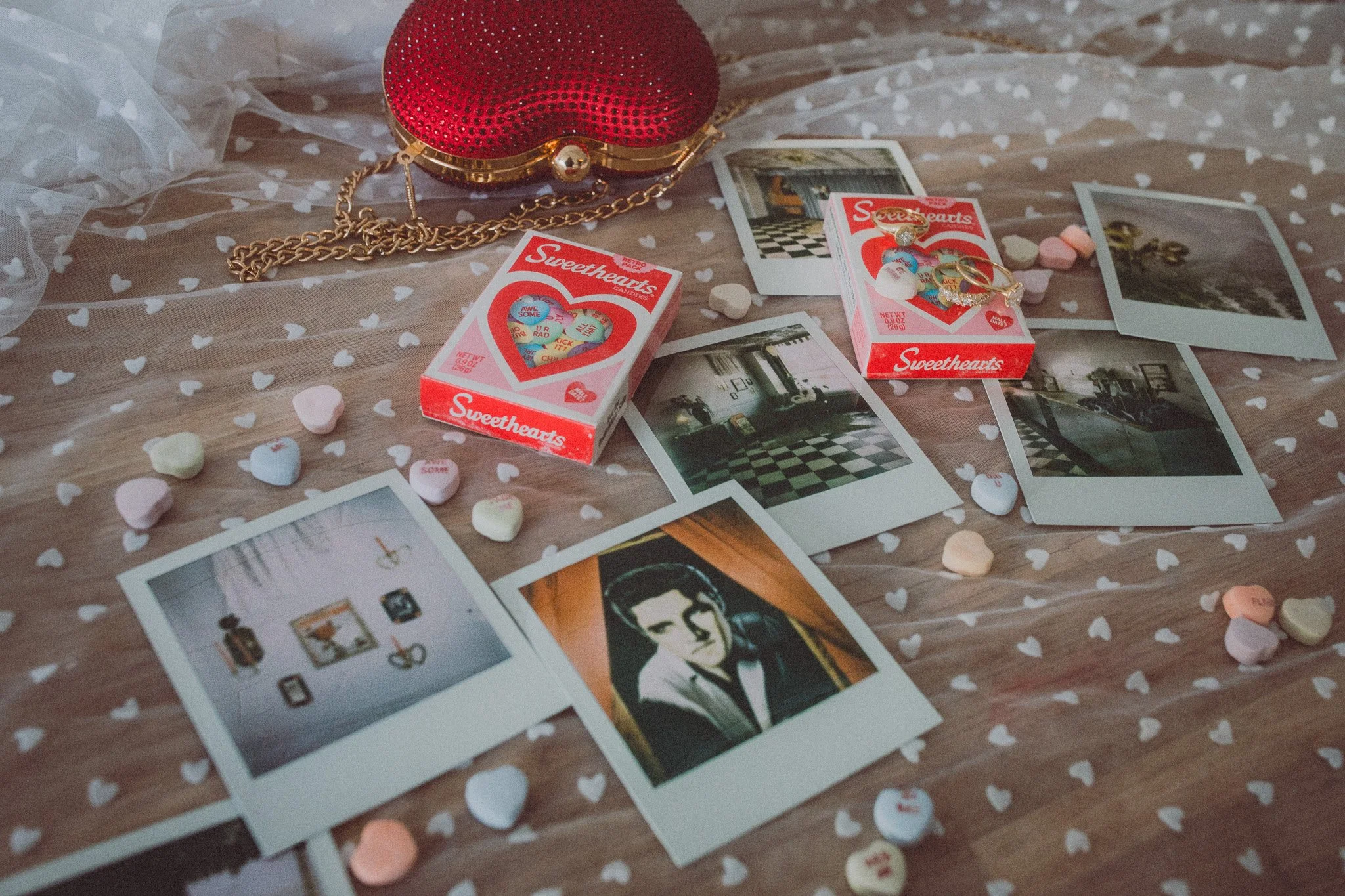 Polaroid photos, a red heart-shaped purse with a gold chain, sweethearts candy hearts, and small jewelry pieces on a table covered with sheer fabric with white heart patterns.