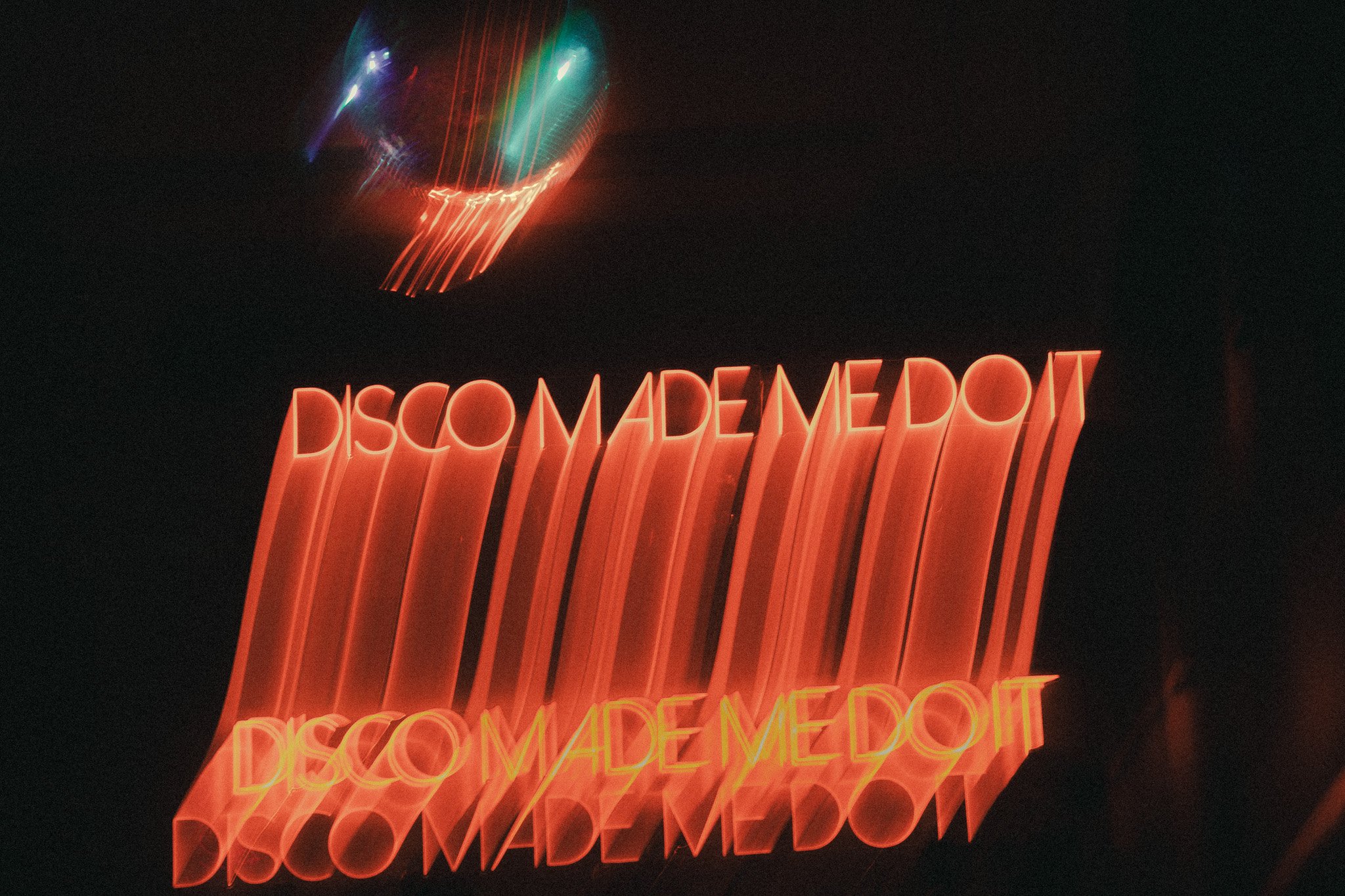 Neon sign displaying the words 'DISCOMADEMDOT' in red, with a reflection below and a dark background, and colorful, blurred lights above.