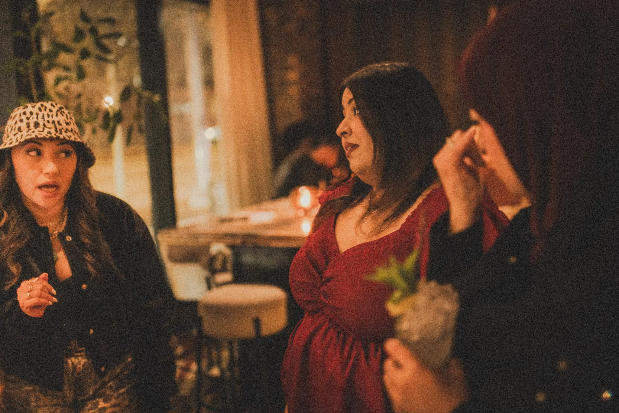 Three women having a conversation in a dimly lit restaurant or bar. One woman on the left is wearing a leopard print hat and black jacket, the woman in the middle is wearing a red dress, and the third woman on the right, partially visible with red ha