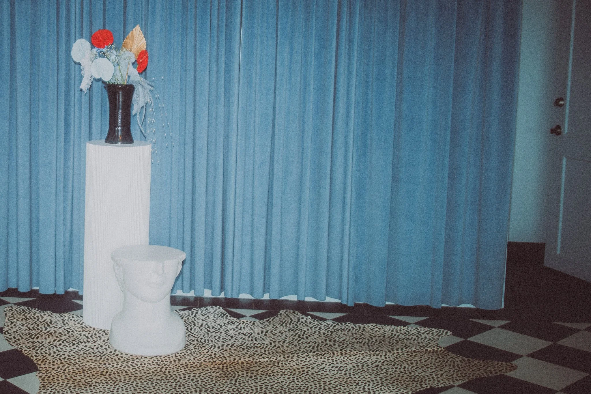 Decorative exhibit with a tall black vase holding a floral arrangement, a white pedestal with a head-shaped sculpture, on a leopard print rug, against blue curtains.