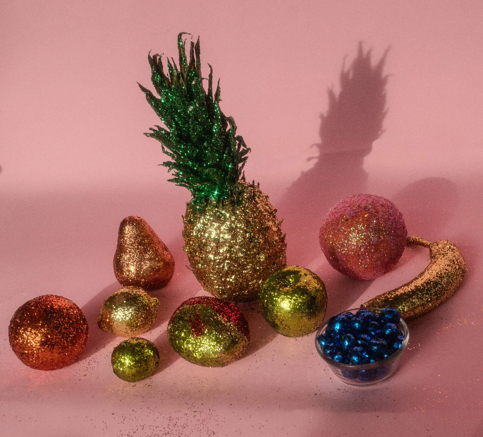 Gift-wrapped fruits including a pineapple, strawberry, lemon, and orange, decorated with glitter and surrounded by a bowl of blue berries, all set on a pink background.