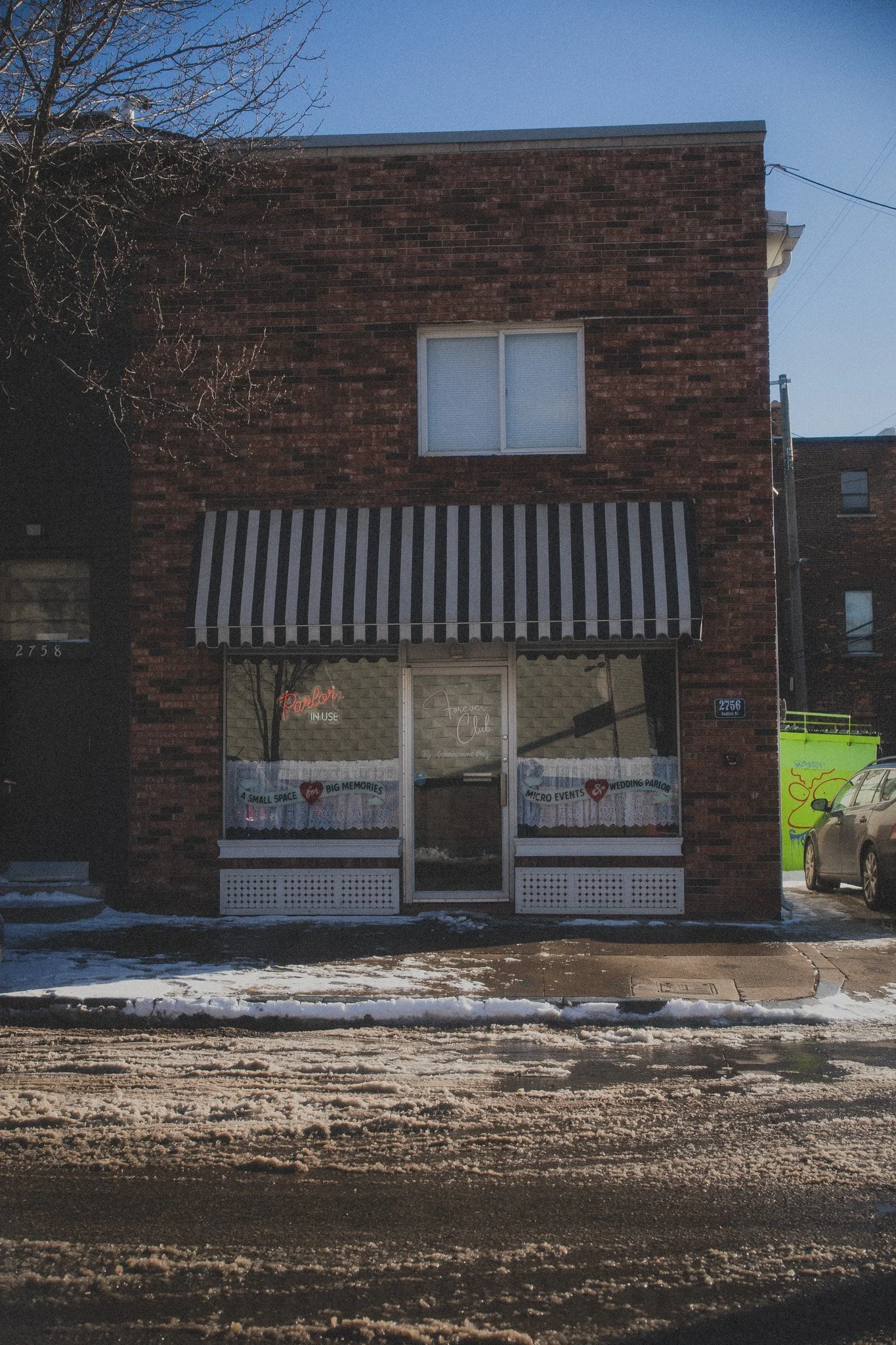 A brick storefront with a striped awning, glass door, and window displaying signs and neon lights. Snow on the ground and a car parked outside.