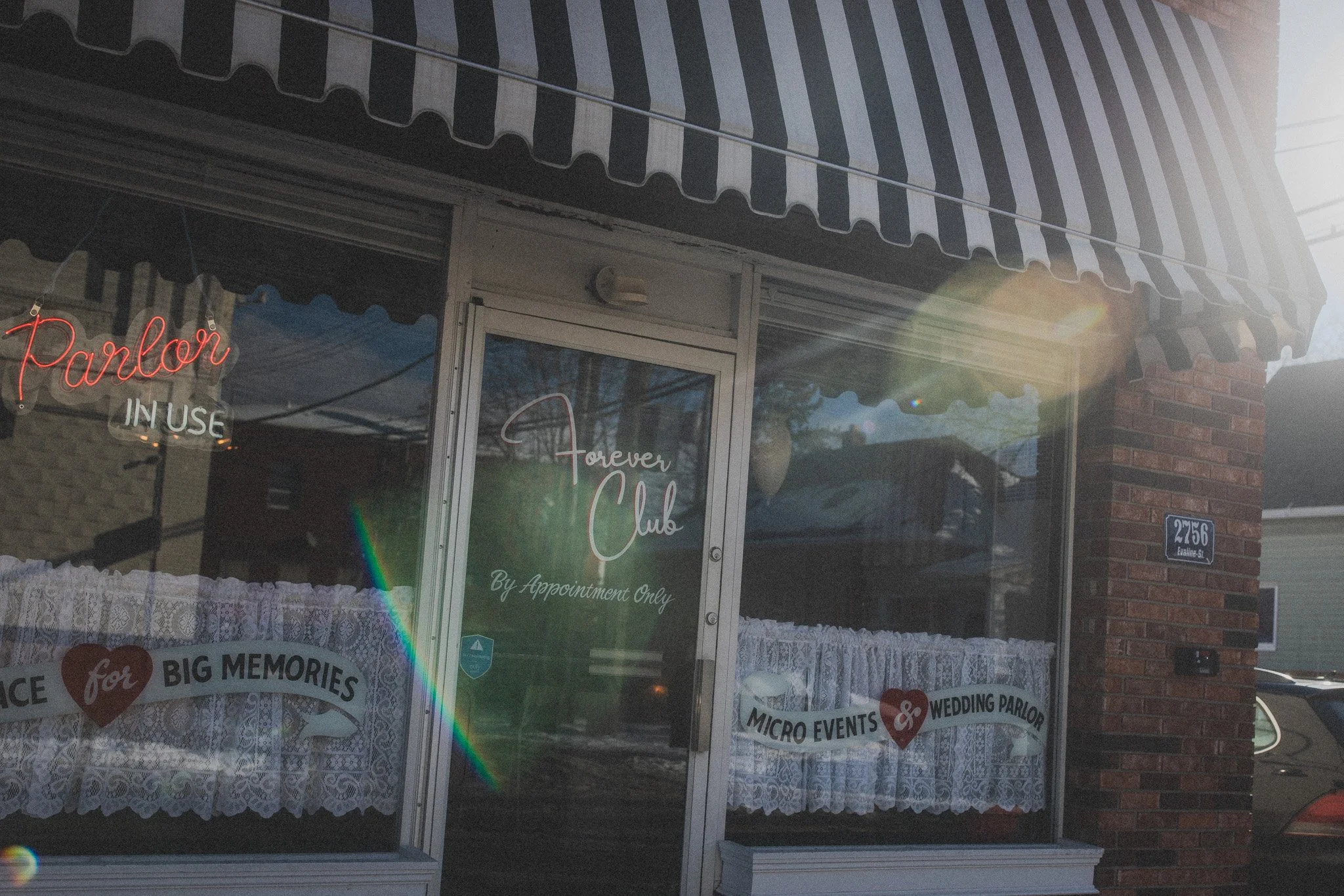  storefront with striped awning, neon sign that says 'Parlor in Use', glass door with 'Forever Club' and 'By Appointment Only' signs, and window decorations that include messages about memories and wedding parlor.