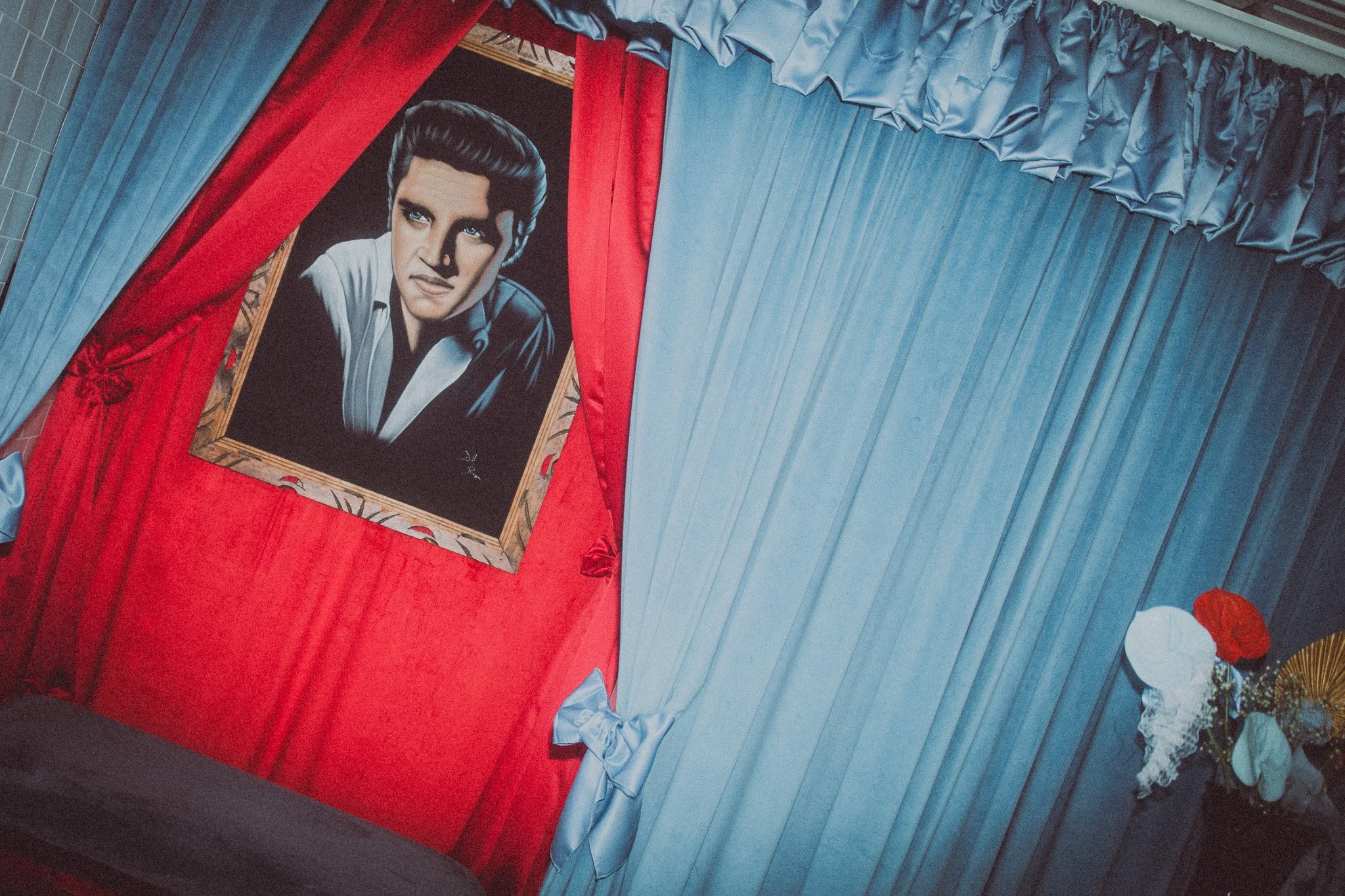 Portrait of Elvis Presley in a black and white shirt with a white collar, framed and displayed on a wall with red and blue curtains.