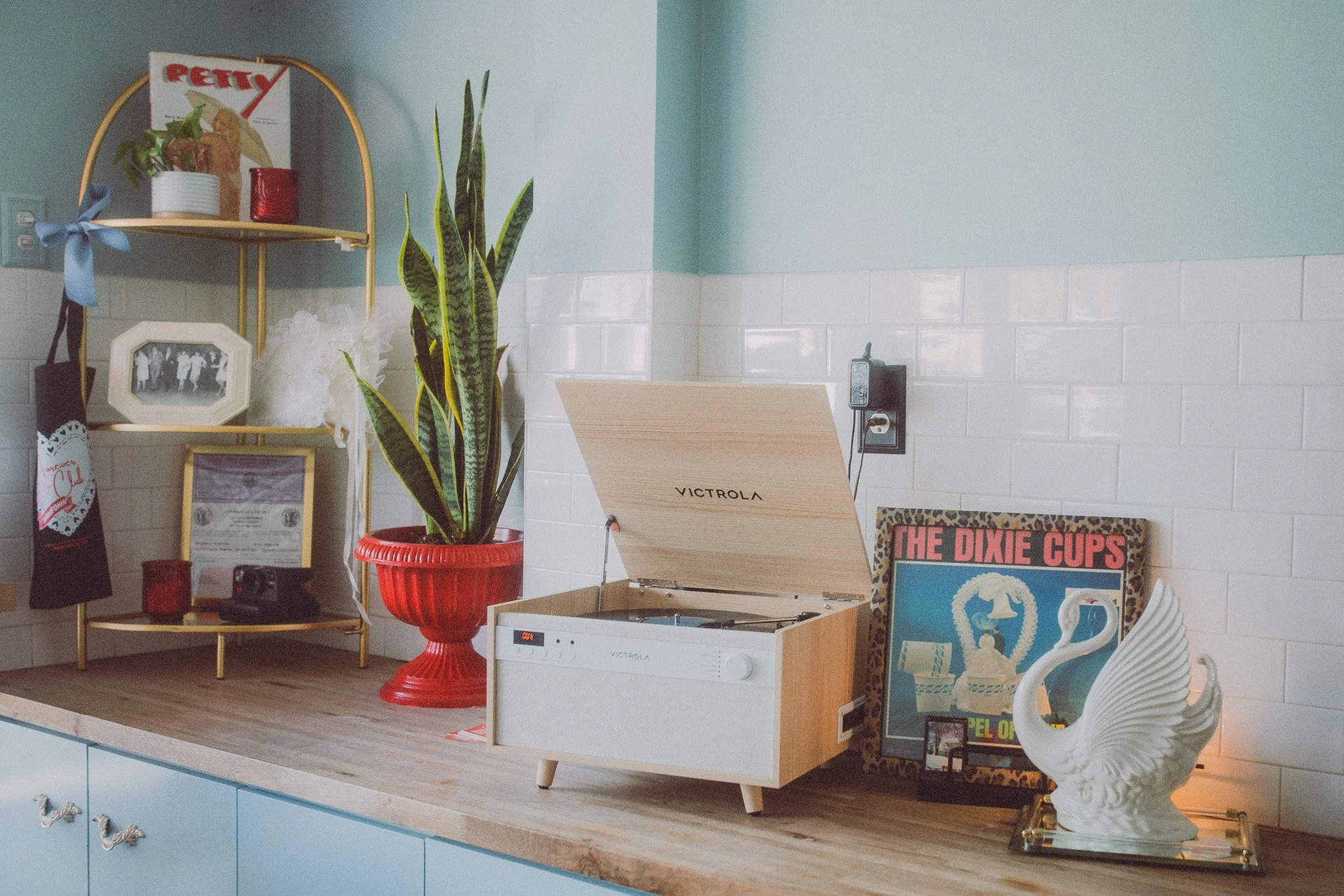 Decorative items arranged on a kitchen counter including a snake plant in a red pot, a vintage record player, a framed poster titled 'The Dixie Cups', a swan-shaped ceramic sculpture, and various small decorative objects and photo frames.