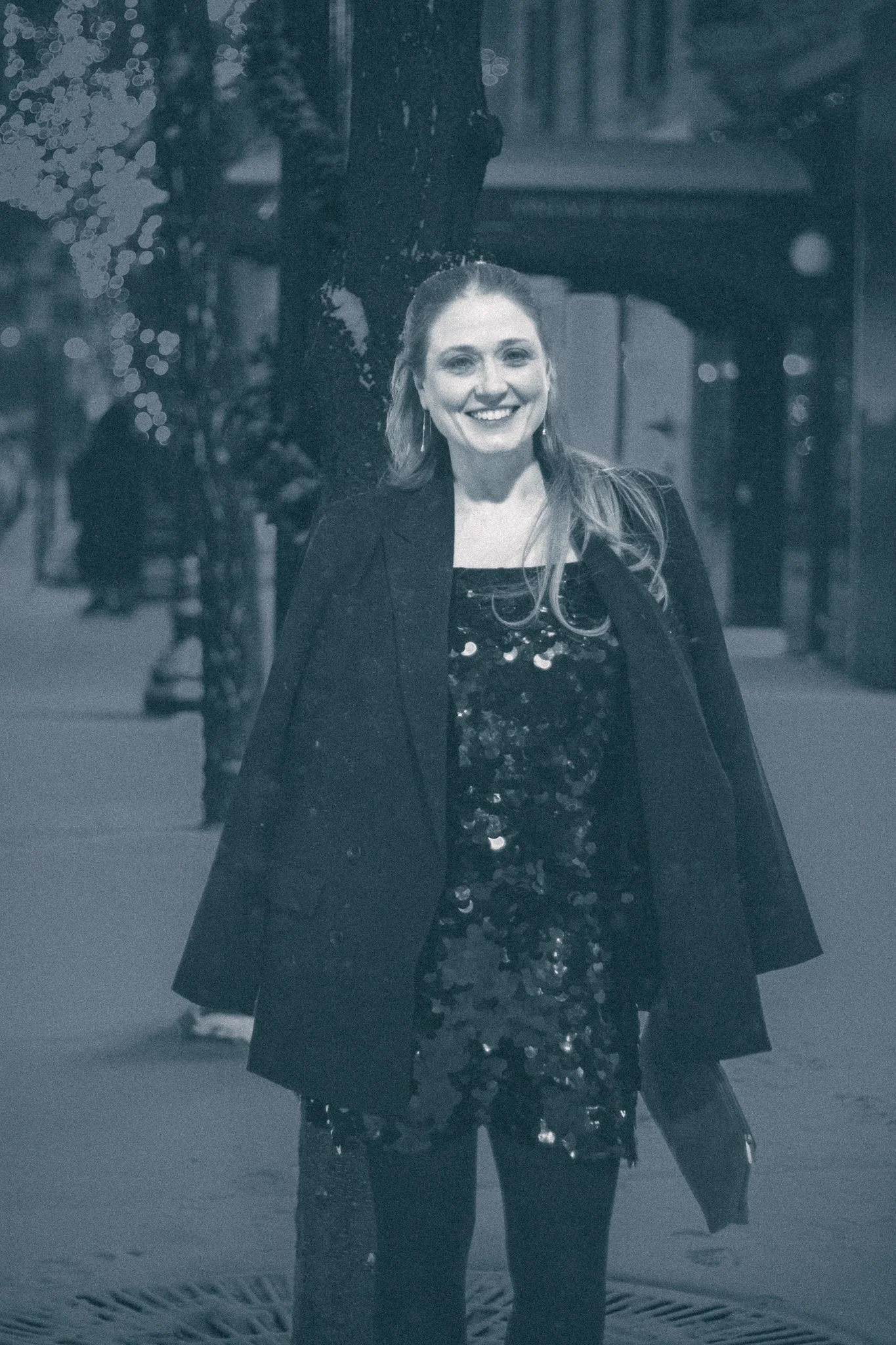 A smiling woman with long hair standing outdoors at night, wearing a black blazer over a sparkly dress, with trees and street lights in the background.