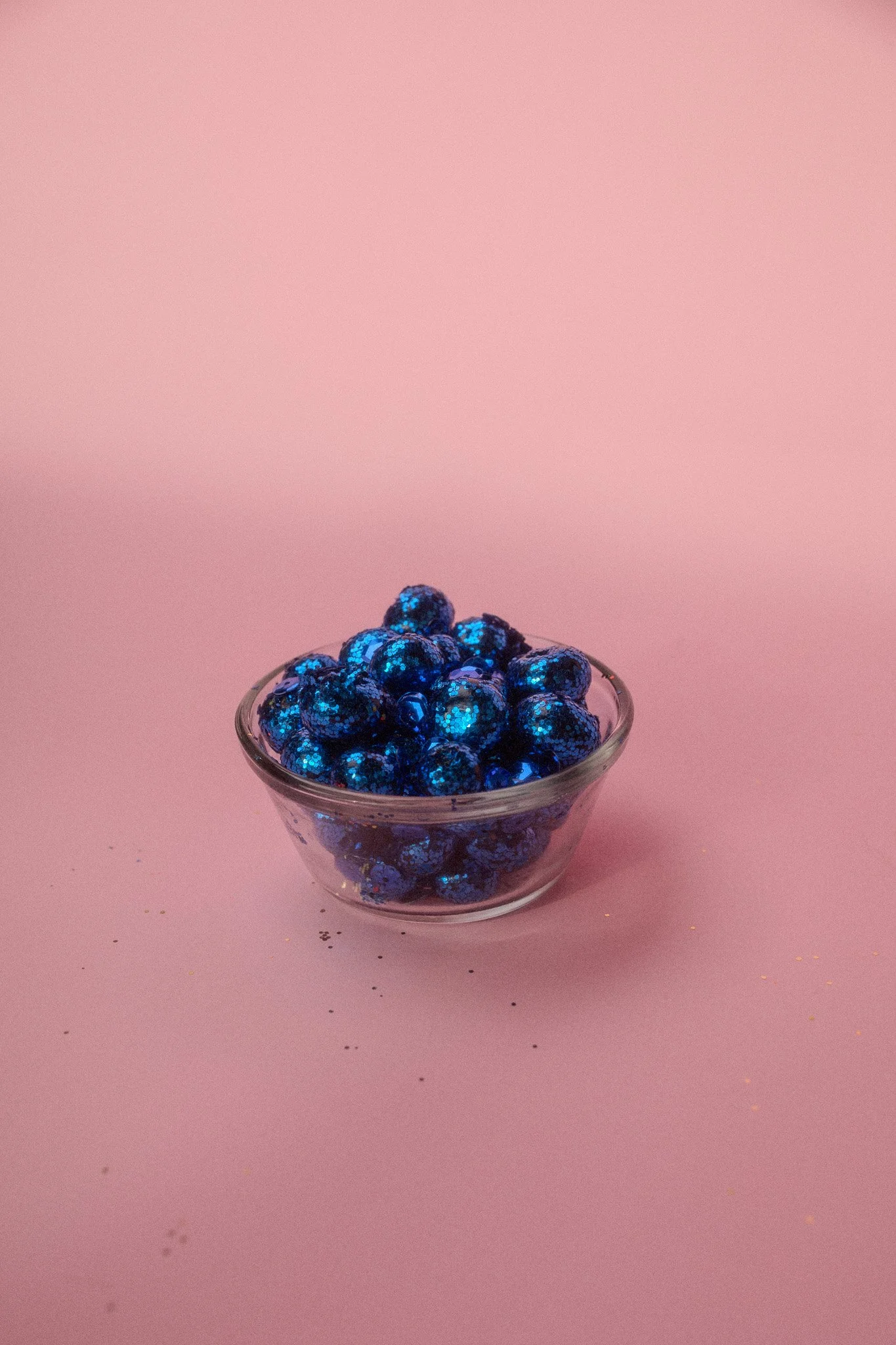 Small glass bowl filled with shiny, dark blue candies or blueberries on pink background.