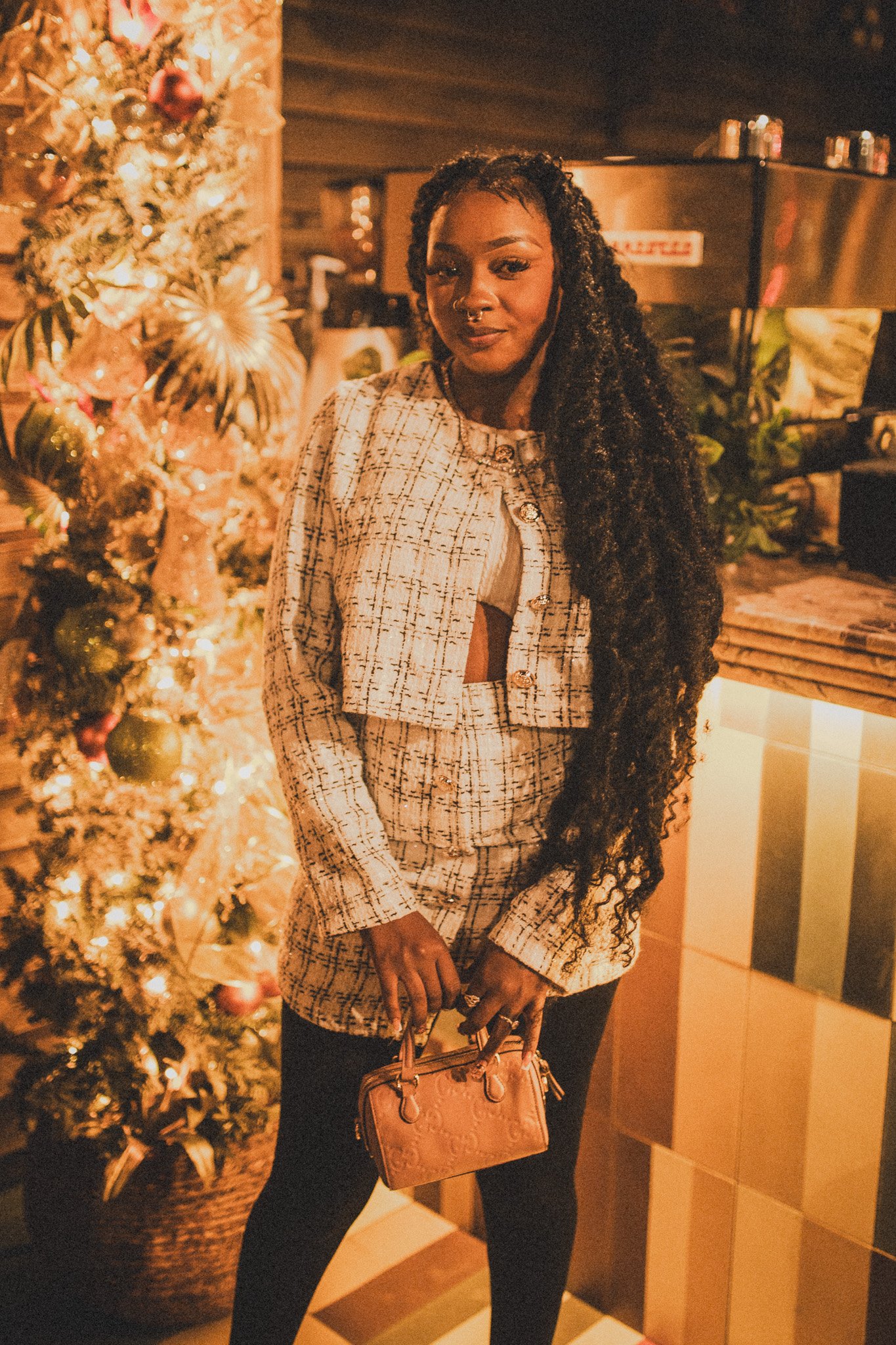 A woman with long, curly hair holding a small pink handbag, standing near a decorated Christmas tree with lights and ornaments, in a warmly lit indoor setting.