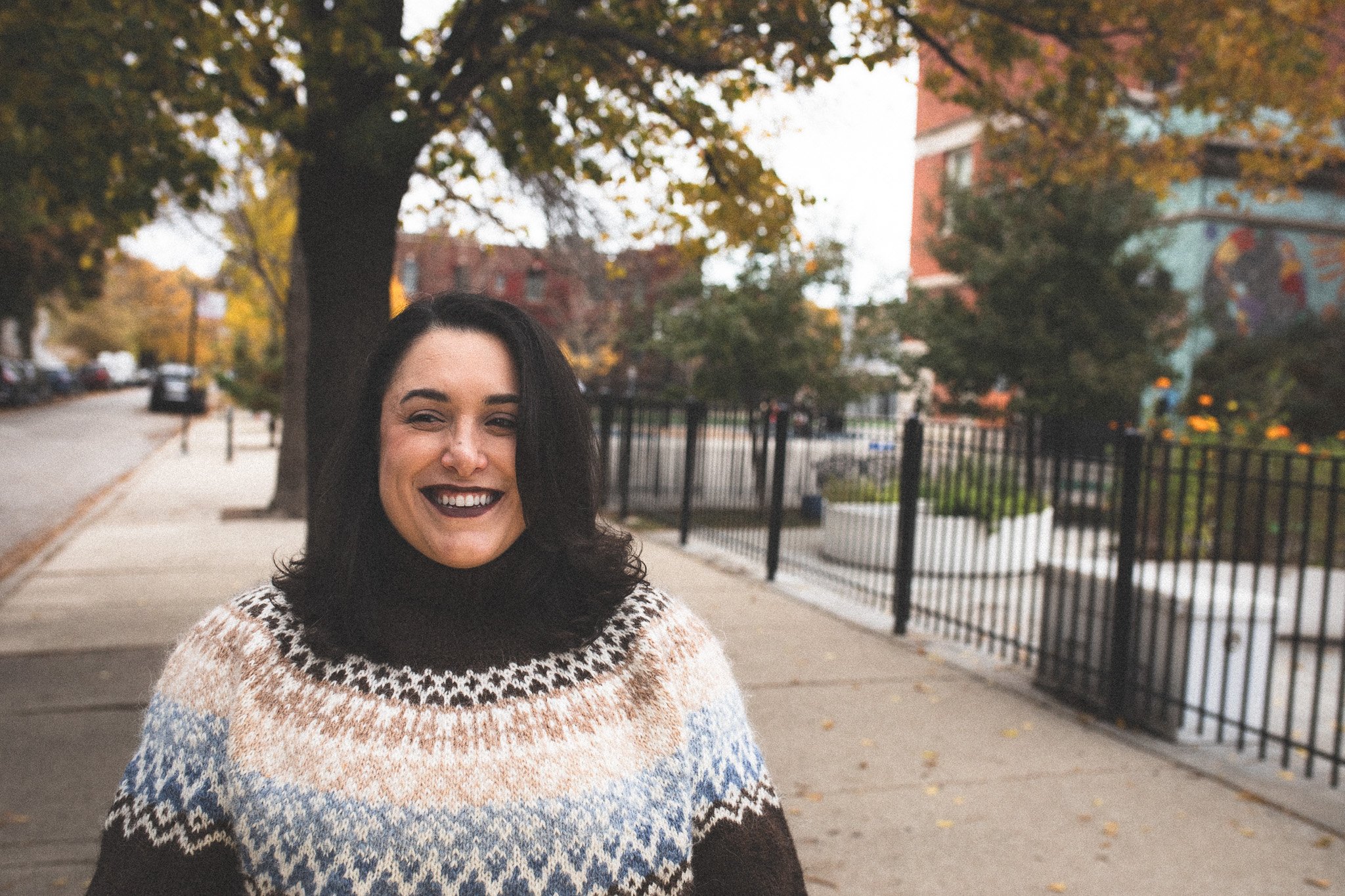 A woman with dark hair smiling on a city sidewalk in front of a tree with fall foliage.