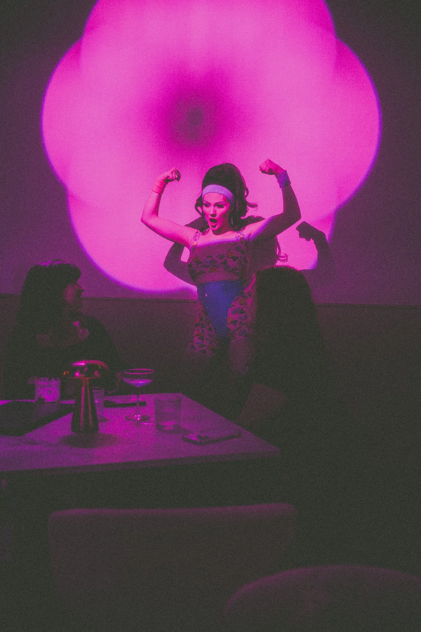 A woman with dark hair in a high ponytail, wearing a leopard print top, strikes a dance pose with flexed arms in front of a pink-lit background and appears surprised. Seated women and drinks are visible in the foreground.