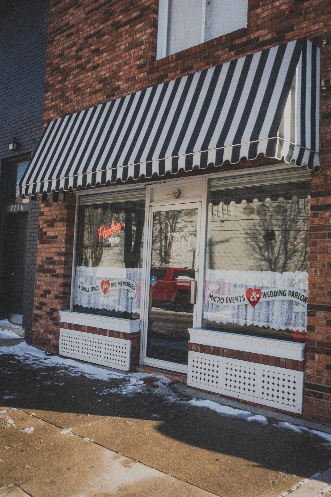  storefront with black and white striped awning, window displays with lace curtains, and signs indicating a wedding parlor and micro events venue, with some snow on the sidewalk.