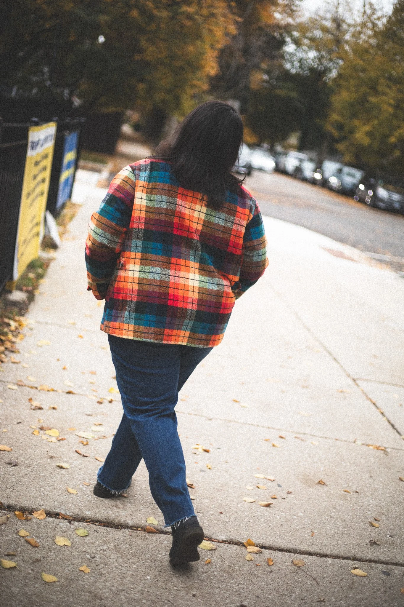 A person with shoulder-length dark hair walking on a sidewalk wearing a multicolored plaid jacket and blue jeans, with autumn trees and parked cars in the background.