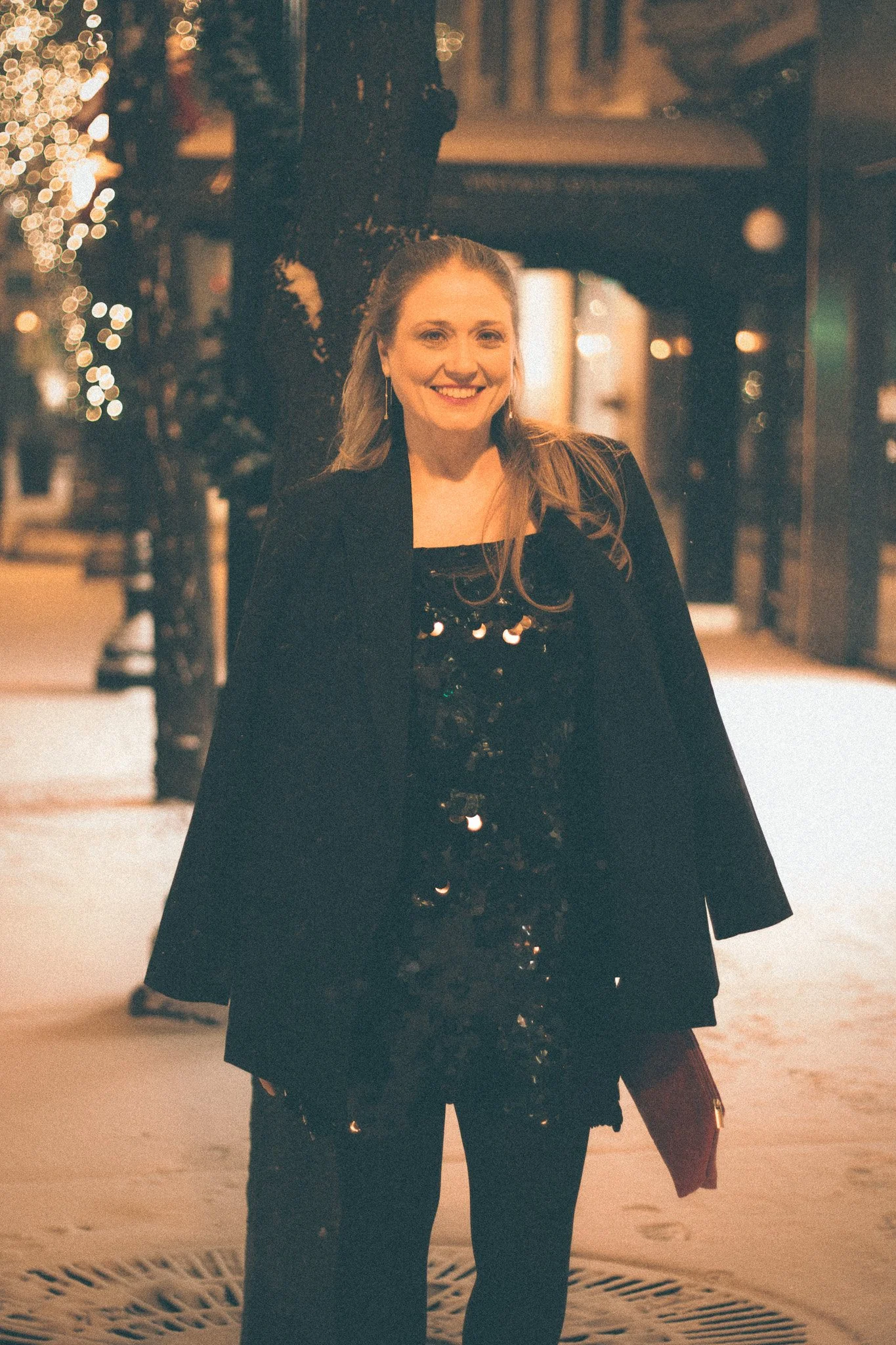 A woman smiling and posing outdoors at night with festive lights in the background.