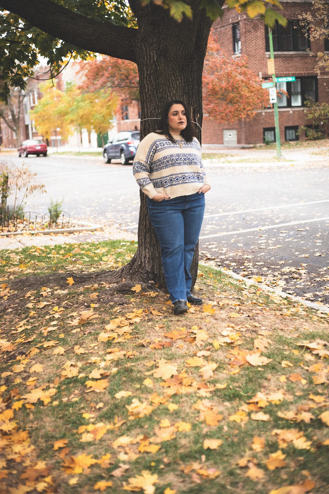 A woman with dark hair, wearing a cream sweater with blue patterns, blue jeans, and black shoes, stands with her hands in her pockets, leaning against a large tree in a park during autumn. Leaves are scattered on the ground, and colorful fall foliage
