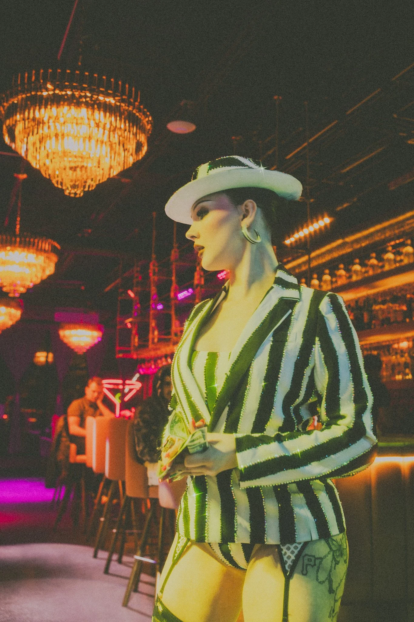 A woman dressed in a striped blazer with matching bold patterned shorts, wearing a white hat, stands in a dimly lit bar with chandeliers and neon signs, with people sitting at the bar in the background.