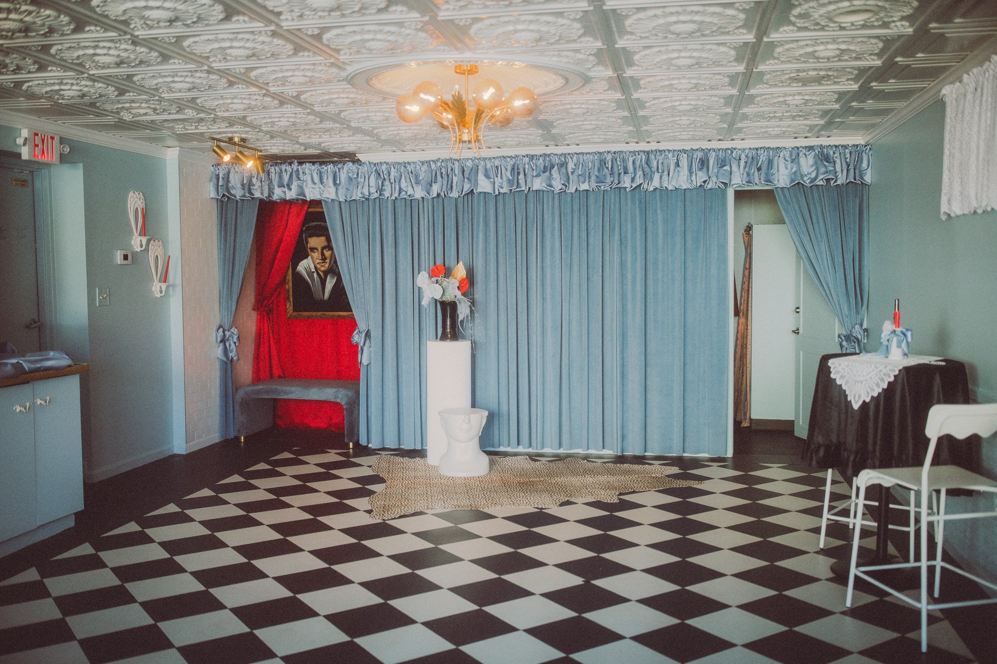 Interior of a room with checkered black and white floor, teal walls, and blue curtains. A portrait of Elvis Presley is partially visible behind the blue curtains on the left, and a white pedestal with a black vase and white and red flowers is in fron